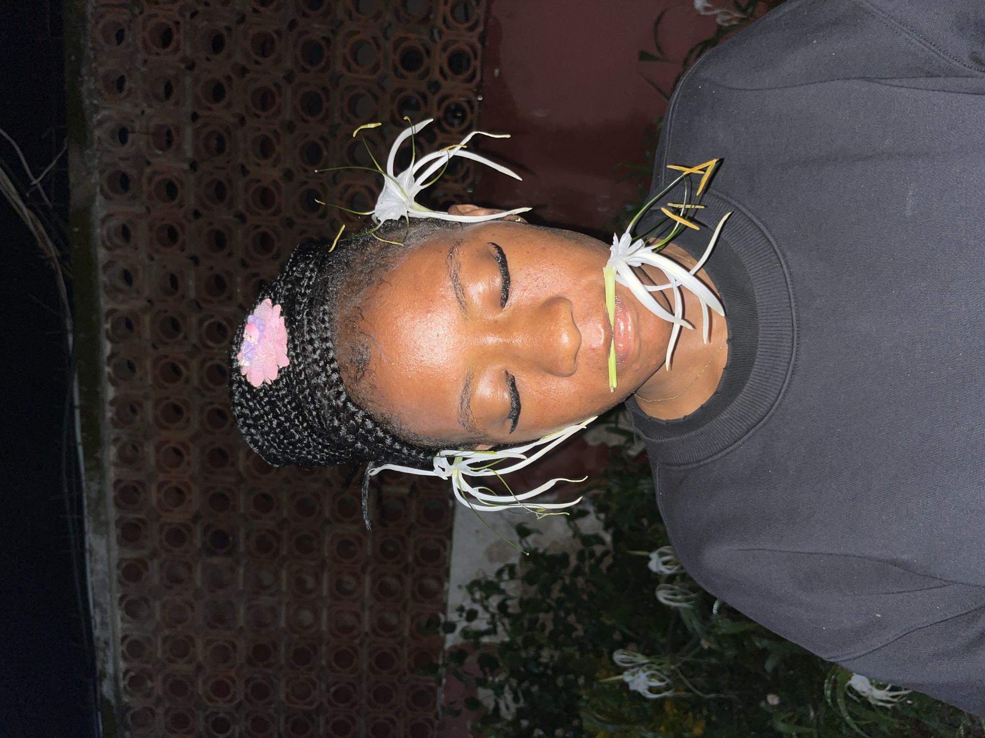 The image shows a close-up of a young Black woman outdoors at night, with a bokeh background of foliage and a textured wall. She is wearing a black sweatshirt. Her eyes are closed, and she has dramatic eyelashes. Her hair is braided and adorned with a small pink flower. She has white spider lilies placed in her hair and one in her mouth, as if she is holding it between her teeth. The overall impression is artistic and surreal. The location is specified as Jos, Nigeria, though no visual cues in the image directly confirm this.