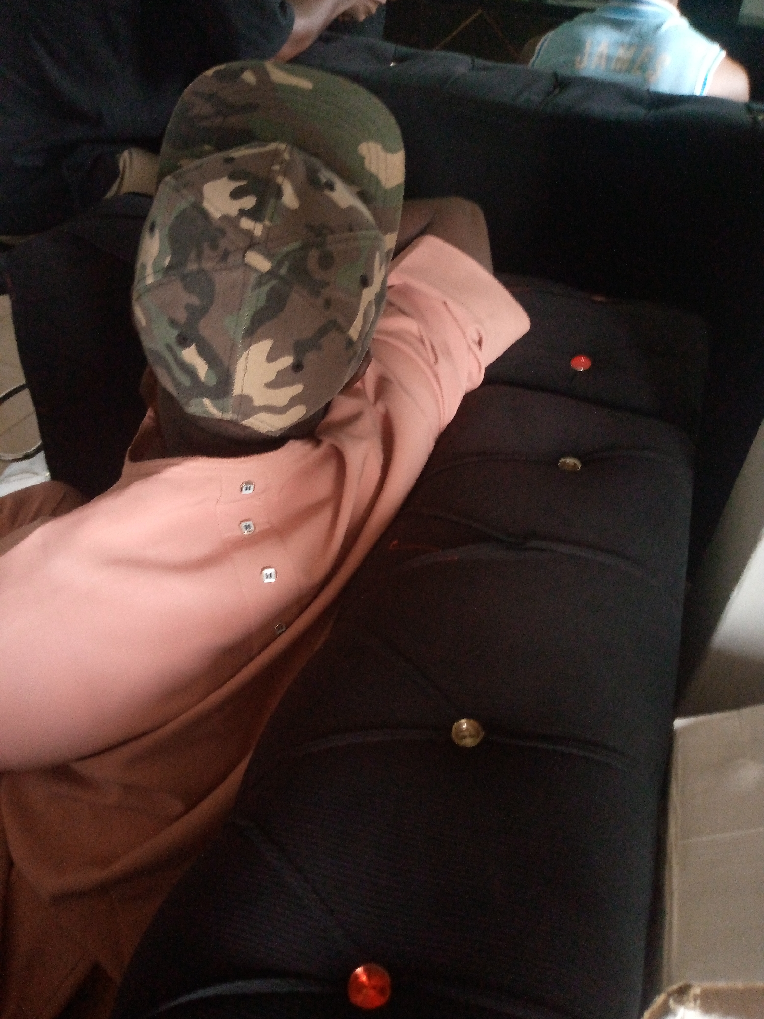A person wearing a camouflage baseball cap and a pink long-sleeved shirt is lying on a black sofa. The person's head is resting on the cap, and their body is partially visible. The sofa has a tufted design with several buttons. A red button is visible on the sofa to the right of the person's shoulder. The background is dark and out of focus, but appears to be an indoor setting, possibly a living room. The image does not provide any specific geographical markers that would indicate its location in Jalingo, Nigeria, or any other city.