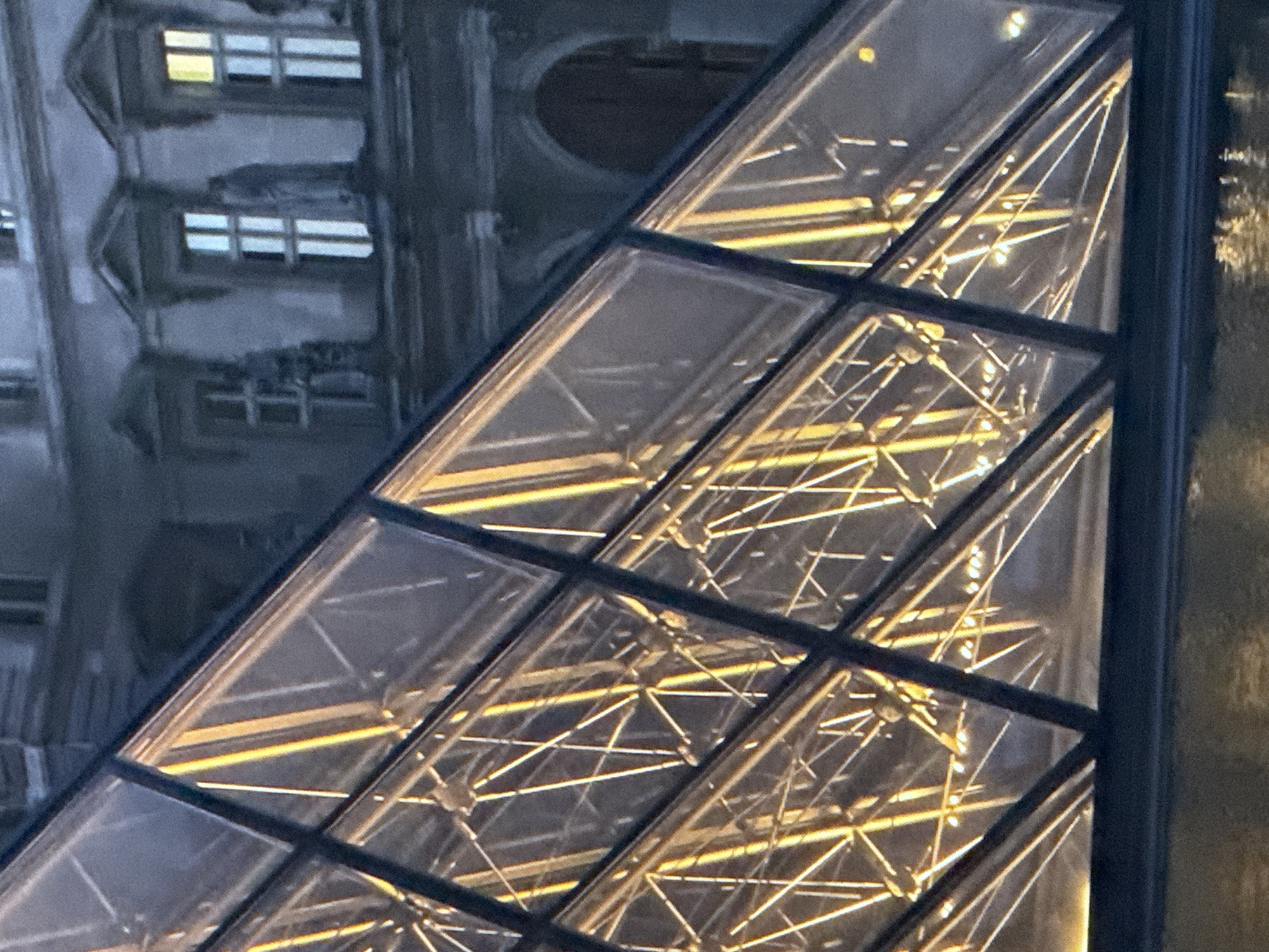 The image shows a close-up view of a glass pyramid structure with illuminated metal framework visible behind the glass panels. The pyramid is angled diagonally across the frame, with its dark metal supports forming geometric patterns. The lighting inside the pyramid is a warm yellow, creating bright vertical and diagonal lines that reflect off the glass. In the background, a classical building with arched windows and decorative elements is visible. The overall scene suggests an architectural landmark, possibly at dusk or night, given the artificial lighting and the subdued tones of the background building.
There is no visual information in the image that allows for the determination of the location being in Saratov, Russia. The architectural style of the building in the background and the glass pyramid are not specific to any particular region without further context.