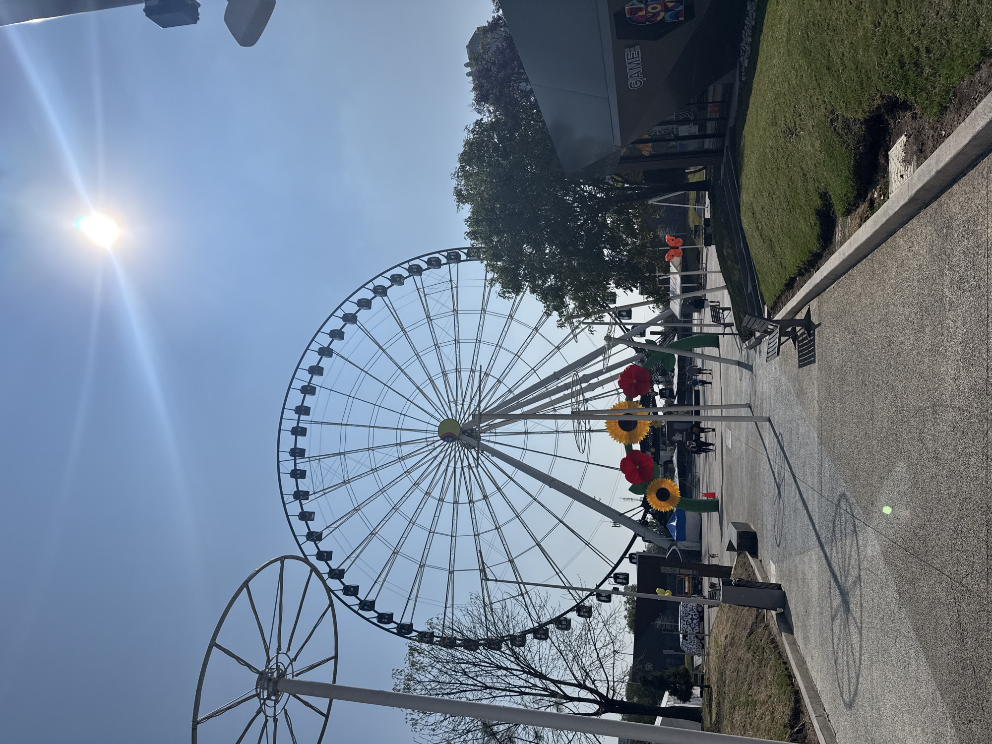 The image shows a large Ferris wheel in Puebla, Mexico, under a bright, clear sky with the sun visible in the upper left. The Ferris wheel is white and features numerous passenger cars. A smaller, partial Ferris wheel structure is visible on the left side of the frame. A paved walkway with park benches and decorative, oversized flowers leads toward the Ferris wheel. Green grass and trees are visible along the walkway. A building with the word "GAME" printed on its side is to the right of the Ferris wheel. The image is rotated, placing the sky and sun on the left.