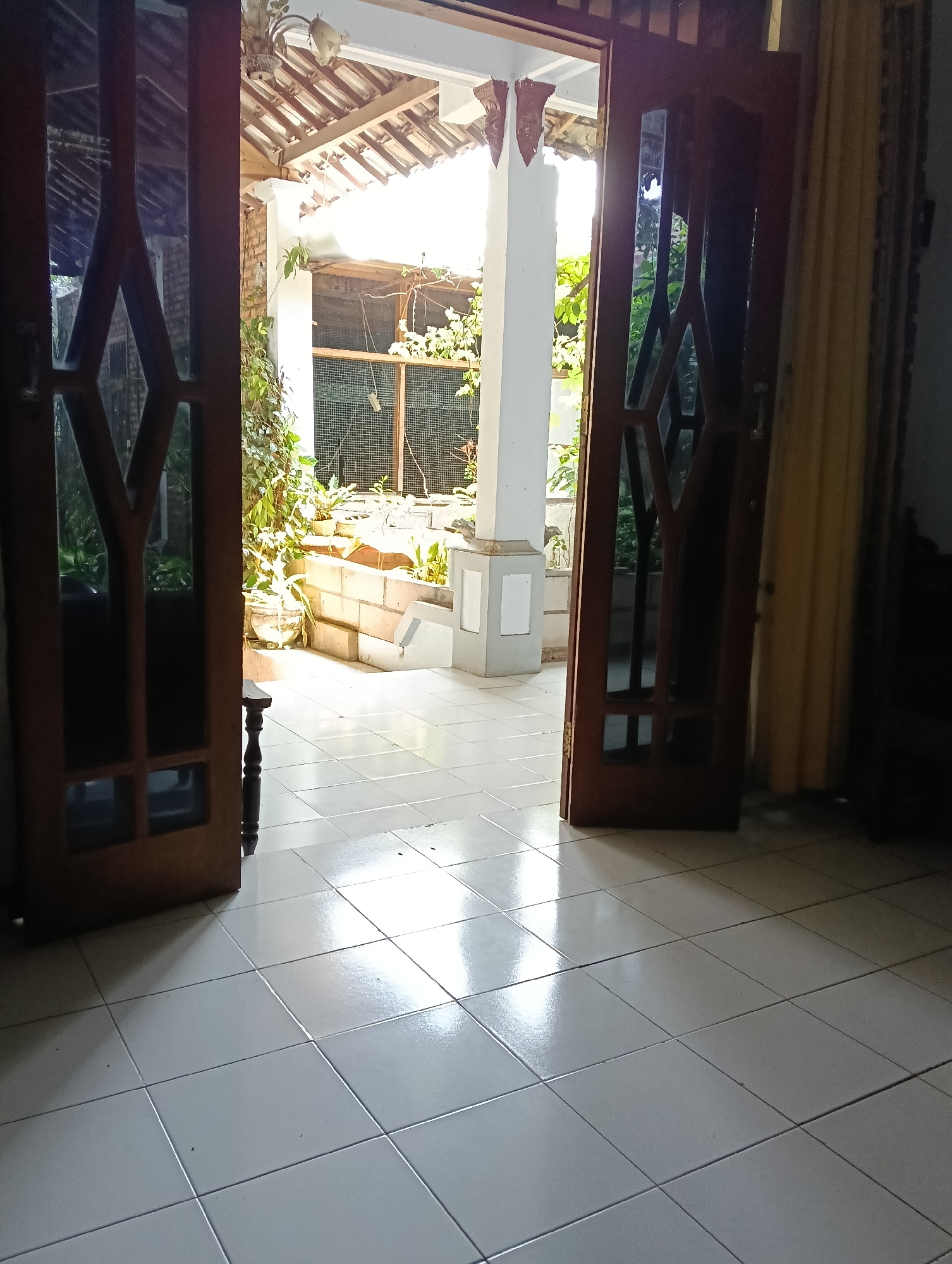 The image shows an open doorway from an indoor tiled floor looking out into an outdoor patio area. Two wooden doors with glass panes are open on either side of the doorway. The patio has a tiled floor, a white pillar, a brick wall with a mesh enclosure, and several potted plants. The area appears to be a residential space in Macanan, Indonesia, likely a backyard or a porch area. The lighting suggests it is daytime, with sunlight filtering in from the outdoor space. No people are visible in the image.