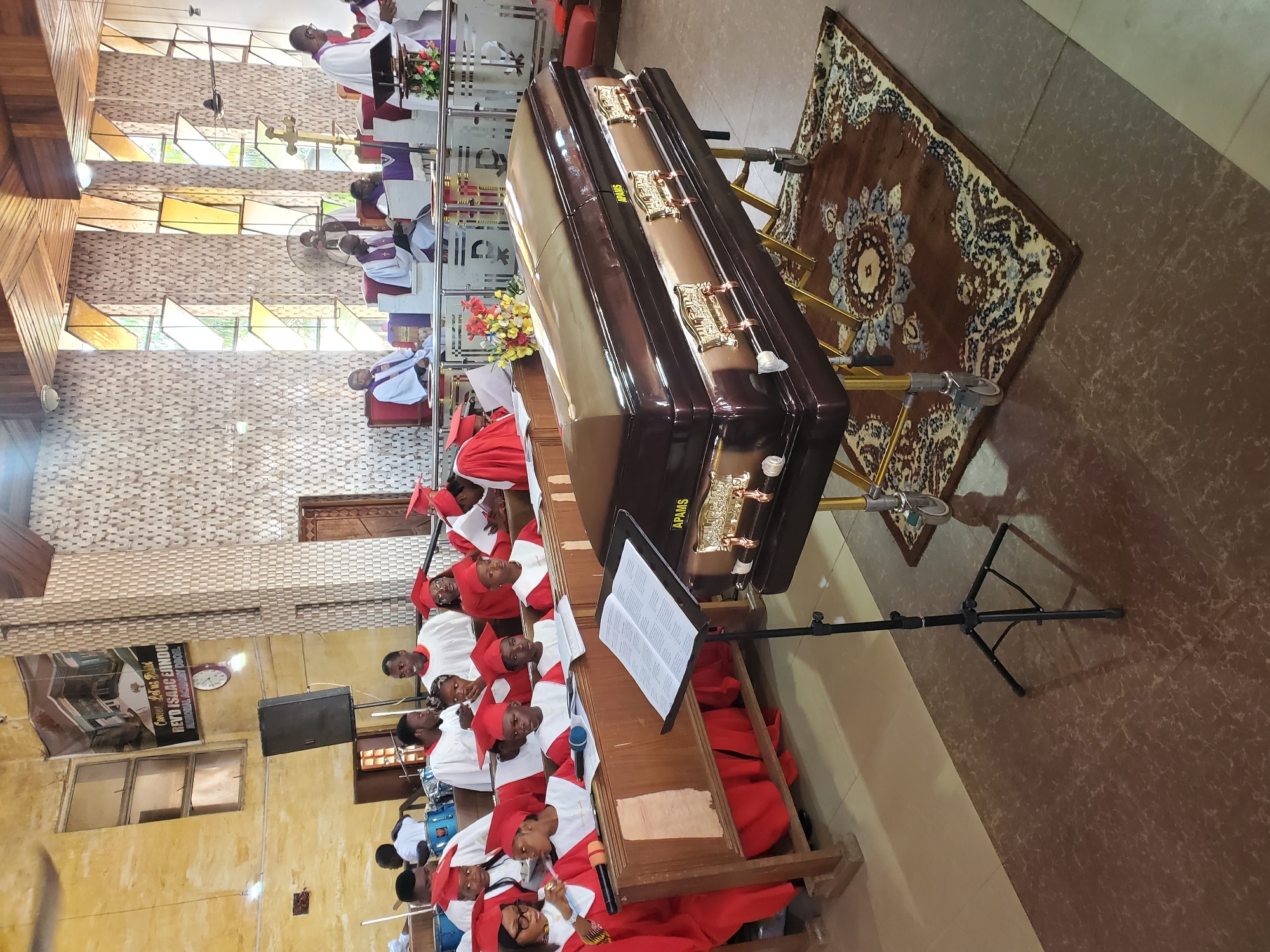 The image captures an indoor funeral service, likely within a church or religious building, observed from a slightly elevated viewpoint. A dark brown, glossy coffin, featuring gold-colored handles and decorative accents, is prominently displayed on a gold-toned stand in the center-right. The word "APAMS" is visible on its side. Beneath the coffin lies a brown rug adorned with a light-colored, ornate floral pattern, set against light brown tiled flooring.
To the left of the coffin, a choir or musical group, comprising adults and children, is seated on wooden benches. They are dressed in white robes complemented by bright red academic caps (mortarboards) and stoles. Microphones on stands are positioned in front of the group, and a black music stand displaying open sheet music is also visible. A blue drum set is partially seen in the lower left corner.
Further back on the left, several individuals, presumably clergy, are seated or standing near an altar or pulpit area, dressed in white robes with purple stoles. Natural light filters through rectangular, patterned windows along the left wall. The interior features light-colored, textured walls and a ceiling with a mix of wooden panels and white sections.
On a wall in the mid-left, a large black speaker is mounted, with a round white wall clock visible above it. A banner or poster on this wall includes text such as "Comm 20th Feb" and "REVD ISAAC EBENU," along with "Divine Peace Gospel." The setting appears solemn and respectful, indicative of a memorial or burial service during daylight hours.