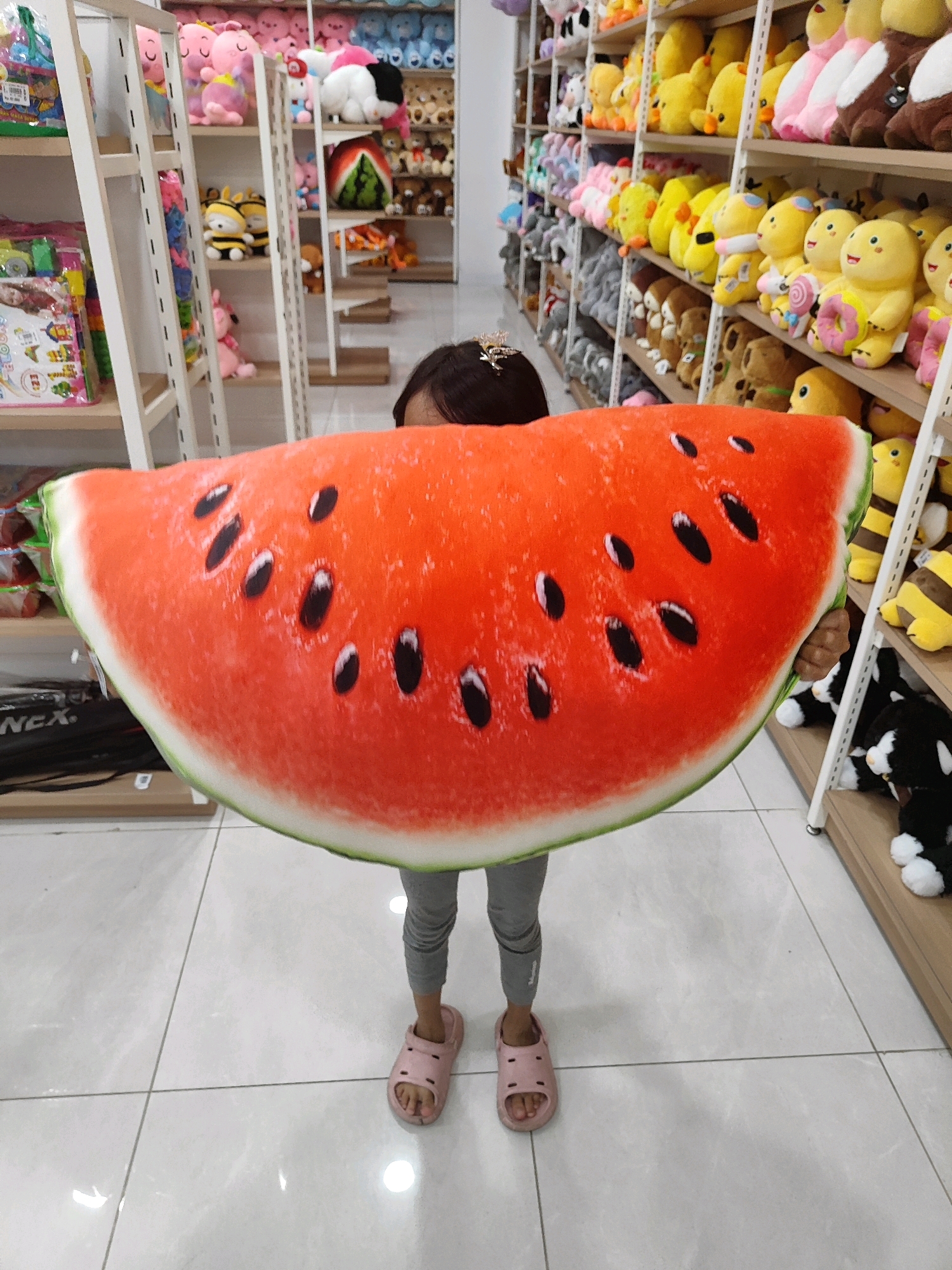 A young child, seen from the chest down, stands in a store filled with stuffed animals. The child is holding a large, round watermelon-shaped pillow. The pillow is red with black seeds and a green rind. The child is wearing grey leggings and pink sandals. The store has white shelving units filled with various stuffed animals, including teddy bears, ducks, and other cartoon characters. The flooring is white tile. The background shows more shelves of stuffed animals, suggesting a toy or gift shop. The scene appears to be indoors, with fluorescent lighting. There is no visible indication of the specific location being Sukowiryo, Indonesia.