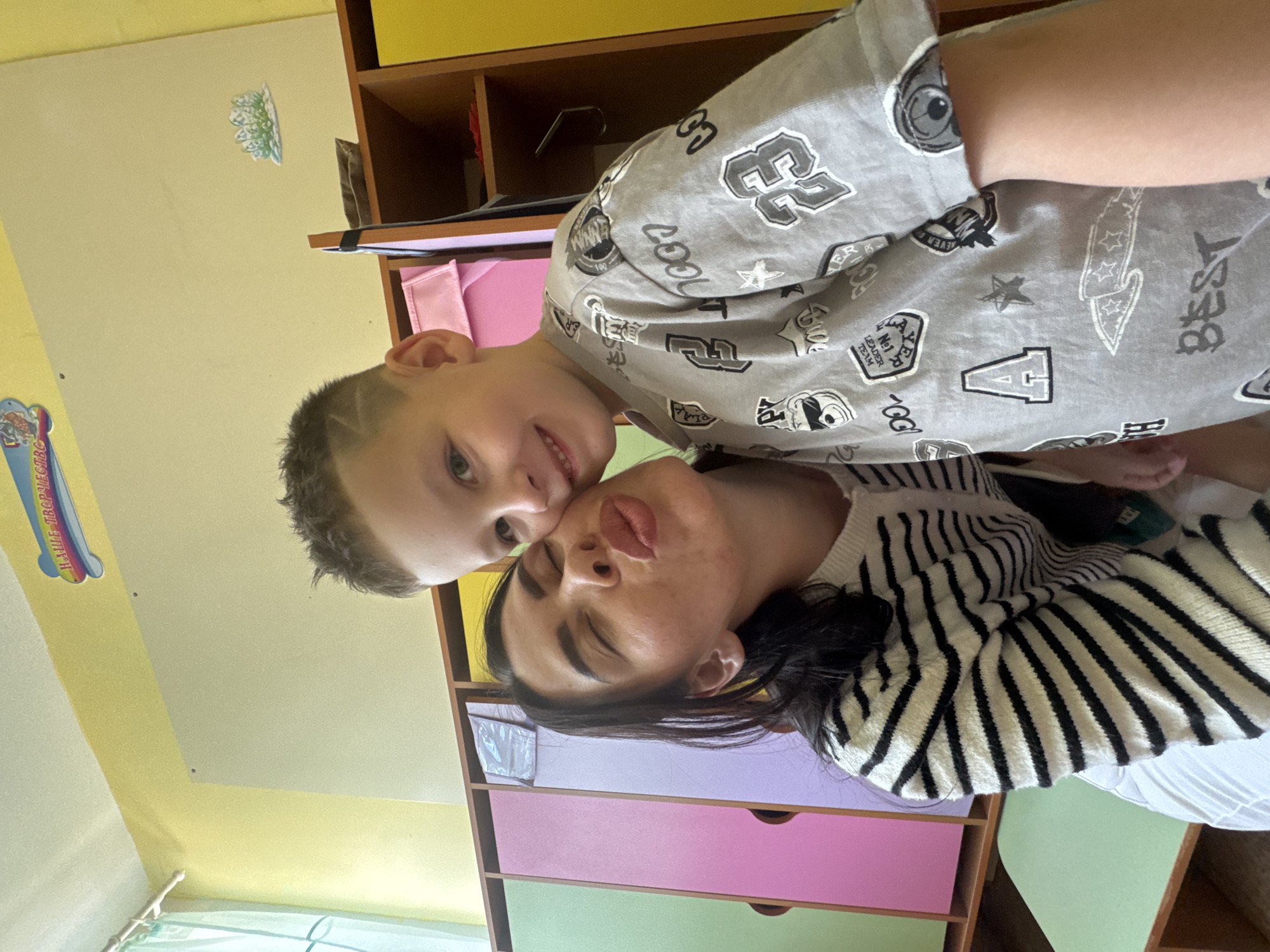 The image shows two people, a boy and a woman, posing for a selfie. The boy is wearing a grey t-shirt with a pattern of numbers, stars, and cartoon-like graphics. He has a haircut with shaved lines on the sides. The woman is wearing a black and white striped sweater. They are positioned close together, with the boy leaning in towards the woman, who is puckering her lips. The background appears to be a room with colorful furniture, including a wooden shelf unit with yellow and pink drawers. There is some text visible on the boy's t-shirt, including "COOL," "BEST," and "LEADER TEAM." A colorful sticker on the wall to the left has text that appears to be in Russian. The overall scene suggests a playful and affectionate moment between the two individuals, likely in a domestic setting. The lighting suggests it is daytime.