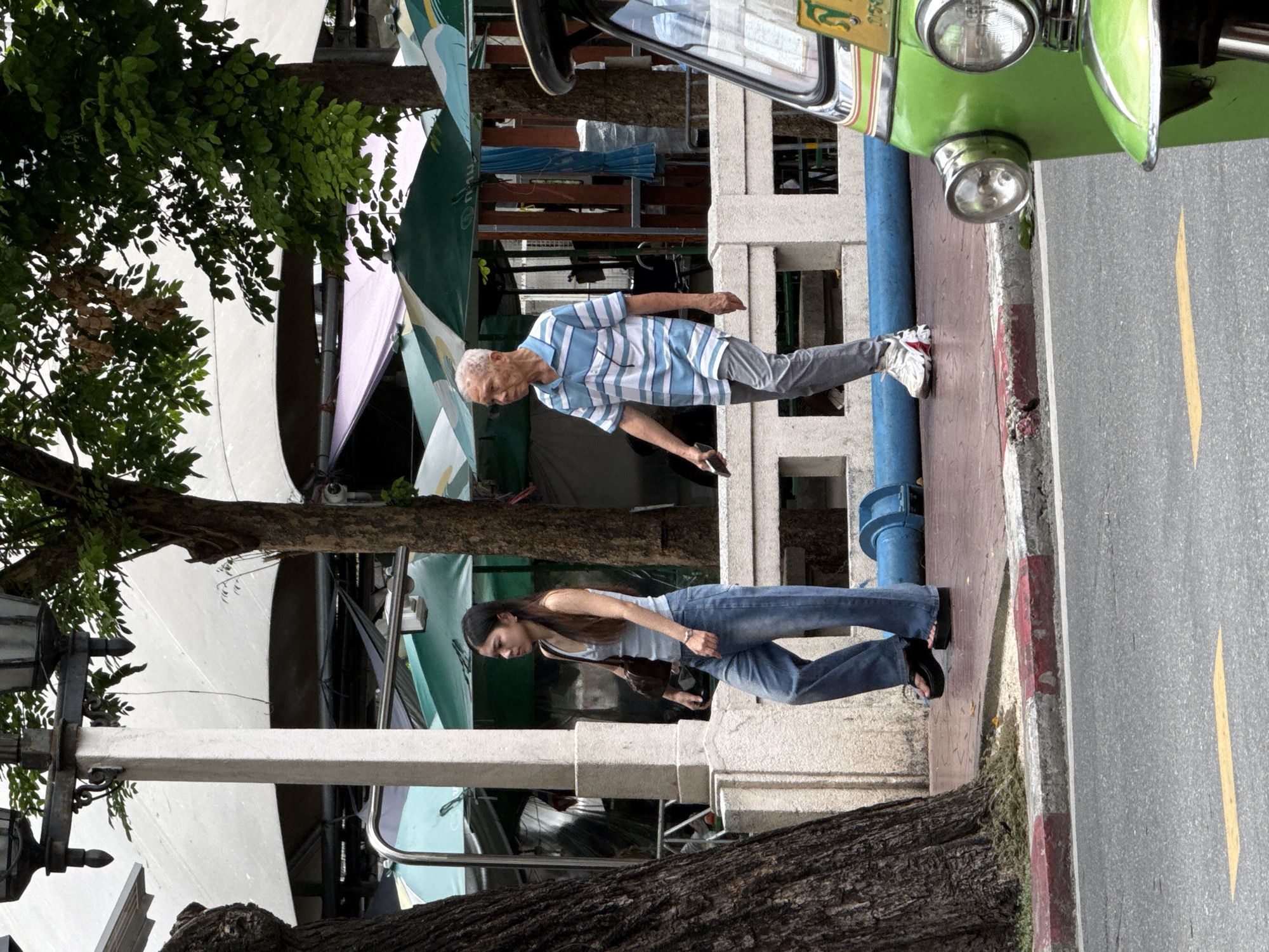 In Bangkok, Thailand, on a street with a gray asphalt road marked with a yellow line, a green tuk-tuk is partially visible on the right. A man with gray hair and a blue and white striped polo shirt walks to the left, holding a dark object in his right hand. A young woman in a light blue tank top and blue jeans walks to the left, carrying a dark brown purse over her left shoulder. They are walking on a red-tiled sidewalk behind a white concrete railing and a large tree trunk on the left. In the background, there are various stalls with green and white awnings.