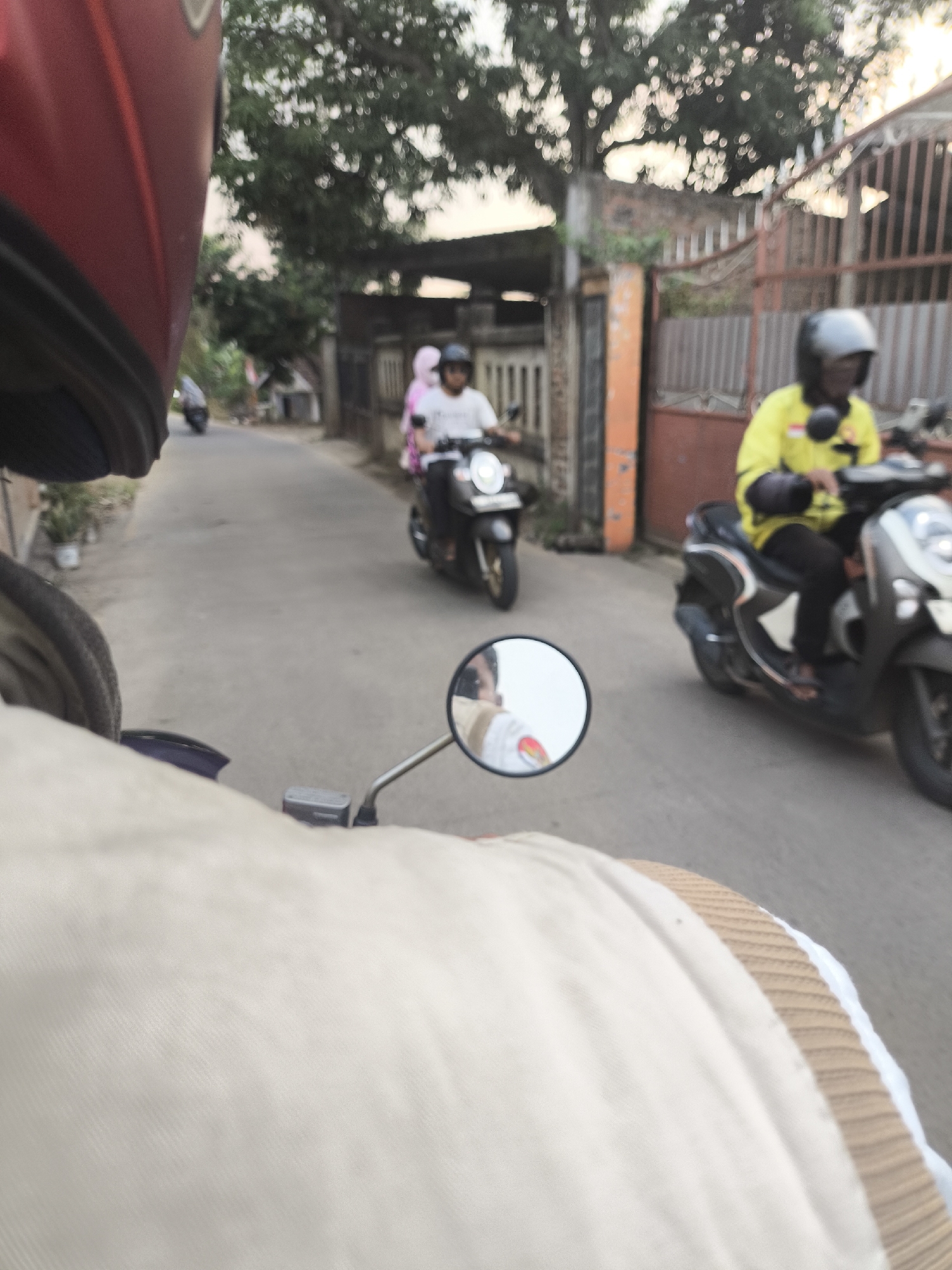 The image captures a street scene in Cilegon, Indonesia, from the perspective of someone riding a motorcycle. In the foreground, the red helmet of the rider and a portion of their light-colored jacket are visible. A motorcycle side mirror reflects the face of the rider and a hint of the person in front of them.
On the road, two other motorcycles are in motion. The motorcycle to the right is ridden by a person wearing a yellow shirt and a black helmet. The motorcycle to the left is ridden by a person wearing a white shirt and a pink headscarf. Both motorcycles are dark-colored scooters.
The environment appears to be a residential street with houses and fences visible on either side. Trees with dense foliage are present in the background, and the lighting suggests it is late afternoon or early evening. The road surface is asphalt. The overall impression is of a casual street scene in a town or city in Indonesia.