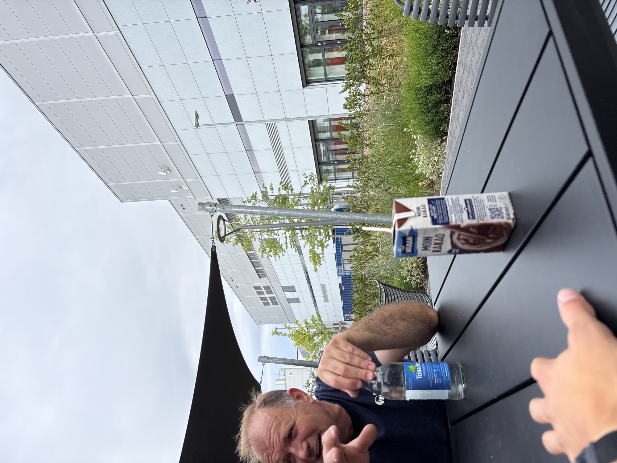 In a close-up, eye-level shot taken outdoors in Mannheim, Germany, a middle-aged man with fair skin and short, graying hair is partially visible from the chest up on the left side of the frame. He is wearing a dark blue t-shirt with a white logo. The man is gesturing with his left hand, which is in focus, and his mouth is open as if speaking. His right hand is also visible, holding a clear plastic water bottle with a blue label that reads "Teinacher MEDIUM".
On a dark gray, slatted outdoor table in the foreground, a carton of chocolate milk from MILRAM, labeled "MOIN KAKAO", is positioned to the right of the water bottle. A second, out-of-focus hand, belonging to the photographer or another person, is visible in the bottom left corner of the frame, reaching towards the table.
In the background, a modern, light-colored building with horizontal paneling and rows of windows dominates the scene. To the left of the building, a black awning provides shade over the area where the man is seated. Green trees and lush vegetation are visible between the table and the building, with some lavender-like plants in the midground. A lamppost stands near the building. The sky is overcast and pale blue. The overall scene suggests a casual outdoor setting, possibly at a cafe or restaurant associated with the building, in Mannheim.