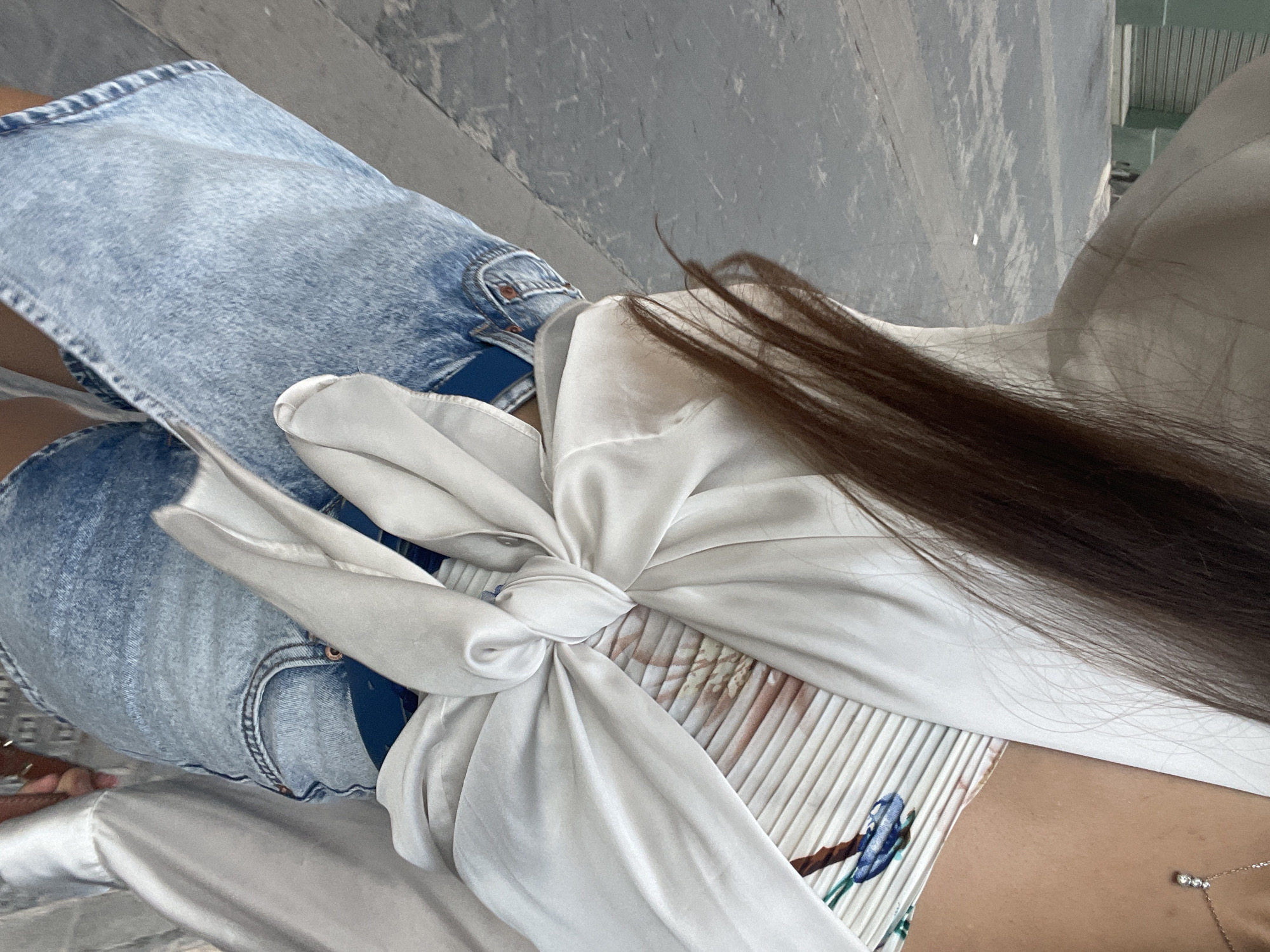 The image shows a woman sitting on steps, possibly in Paiporta, Spain. She is wearing a light wash denim skirt and a white, silky top with a large bow tied at the waist. The top appears to have a pleated section with a floral print visible beneath the bow. A necklace is visible around her neck, and long brown hair rests on her shoulder. The steps are made of concrete or a similar material.