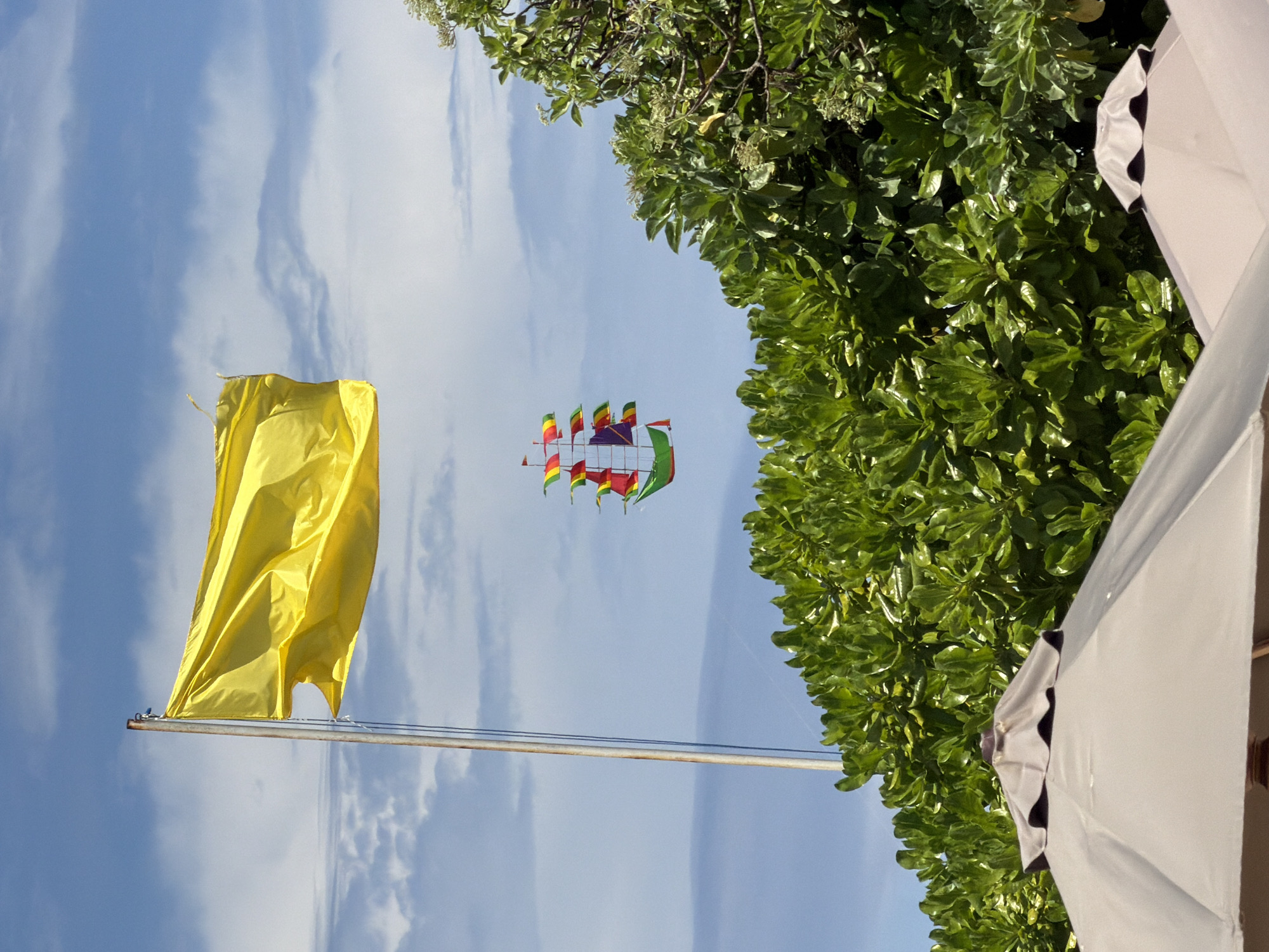 The image shows a partly cloudy sky in Nusa Dua, Indonesia. A yellow flag hangs limp on a metal pole. In the sky, a kite shaped like a ship with colorful flags flies. Lush green foliage is visible to the right, partially obscuring the sky. The edge of a white umbrella is visible on the lower right side.