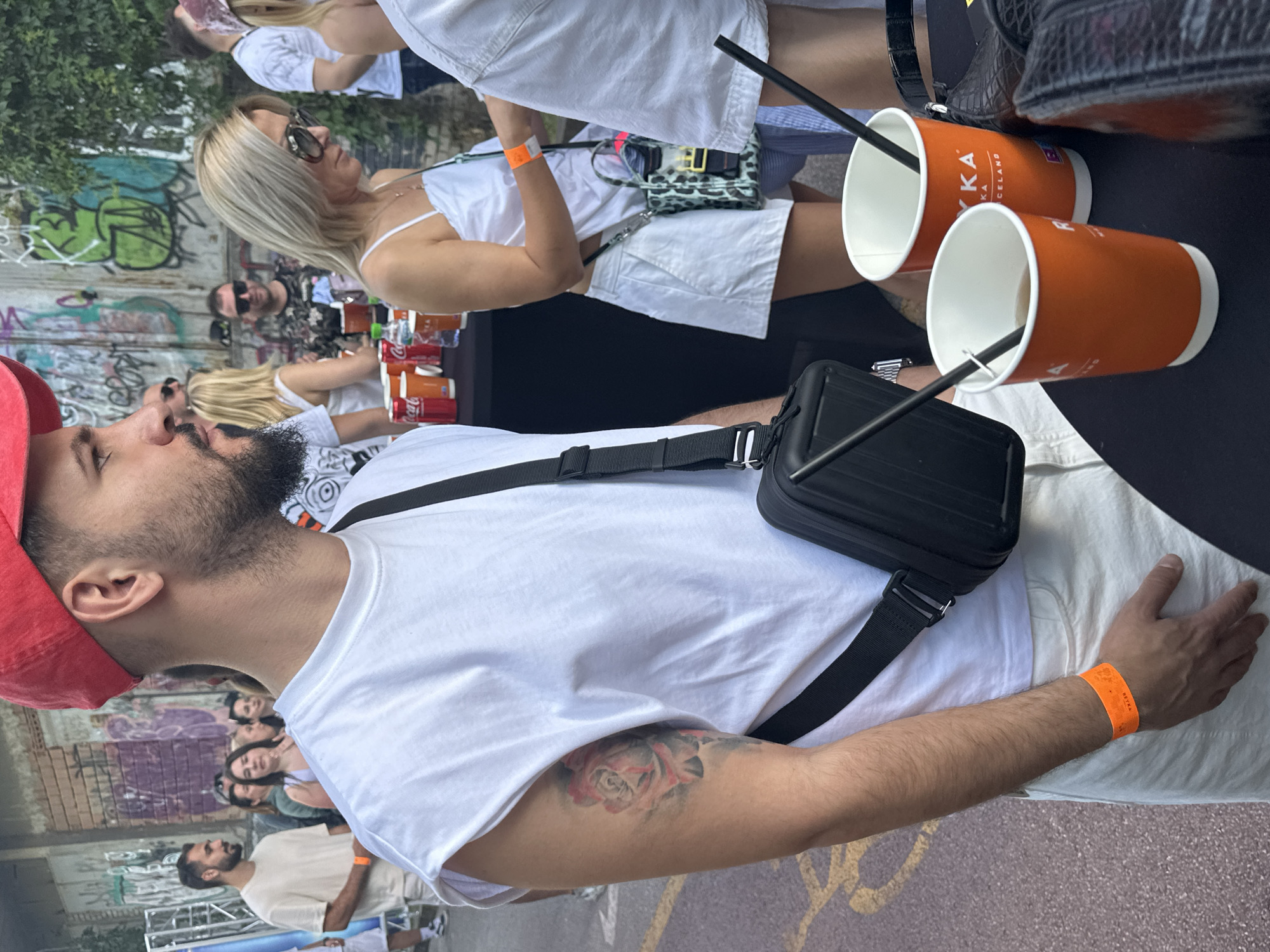 The image shows a street scene in Plovdiv, Bulgaria. The perspective is from above, looking down. A man wearing a red cap and a white T-shirt with a black shoulder bag is in the foreground, facing left. He has a beard and a tattoo on his upper arm. To his right, on a black surface, are two orange cups with black straws. In the background, a woman with blonde hair and sunglasses is wearing a white dress and a patterned handbag. Other people are visible in the background, also wearing white. Graffiti-covered walls are visible in the background, indicating an urban setting. The ground has faded graffiti as well.