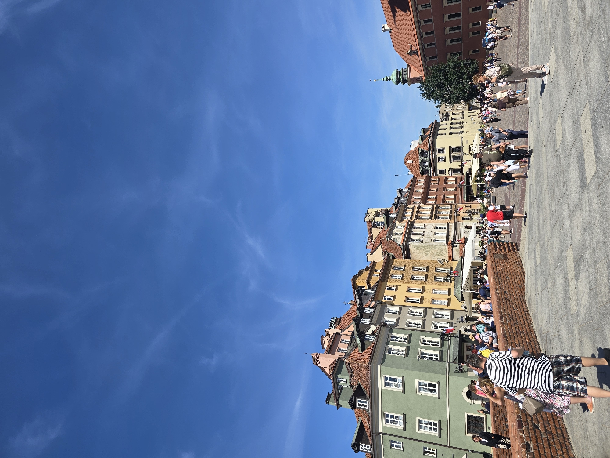 An eye-level, wide-angle shot captures the Old Town Market Square in Warsaw, Poland, on a bright, sunny day. The sky is a clear, vibrant blue with a few wispy white clouds. The square is paved with grey stone tiles and is bustling with people.
In the foreground, a red brick wall separates the paved area from a slightly elevated walkway. Several people are walking and standing on the walkway and in the square. A man in a striped shirt and plaid shorts is facing away from the camera, with a woman in a floral dress walking beside him.
Behind the brick wall, a row of colorful buildings lines the square. These buildings have terracotta-tiled roofs and are painted in various shades of green, pink, yellow, and beige. Many of the buildings feature white-framed windows and dormer windows.
Further into the square, more people are visible, some walking in groups, others sitting at outdoor cafe tables. In the background, a green-domed tower with a spire rises above the buildings, characteristic of Warsaw's Old Town architecture. The overall atmosphere is one of a lively tourist destination.