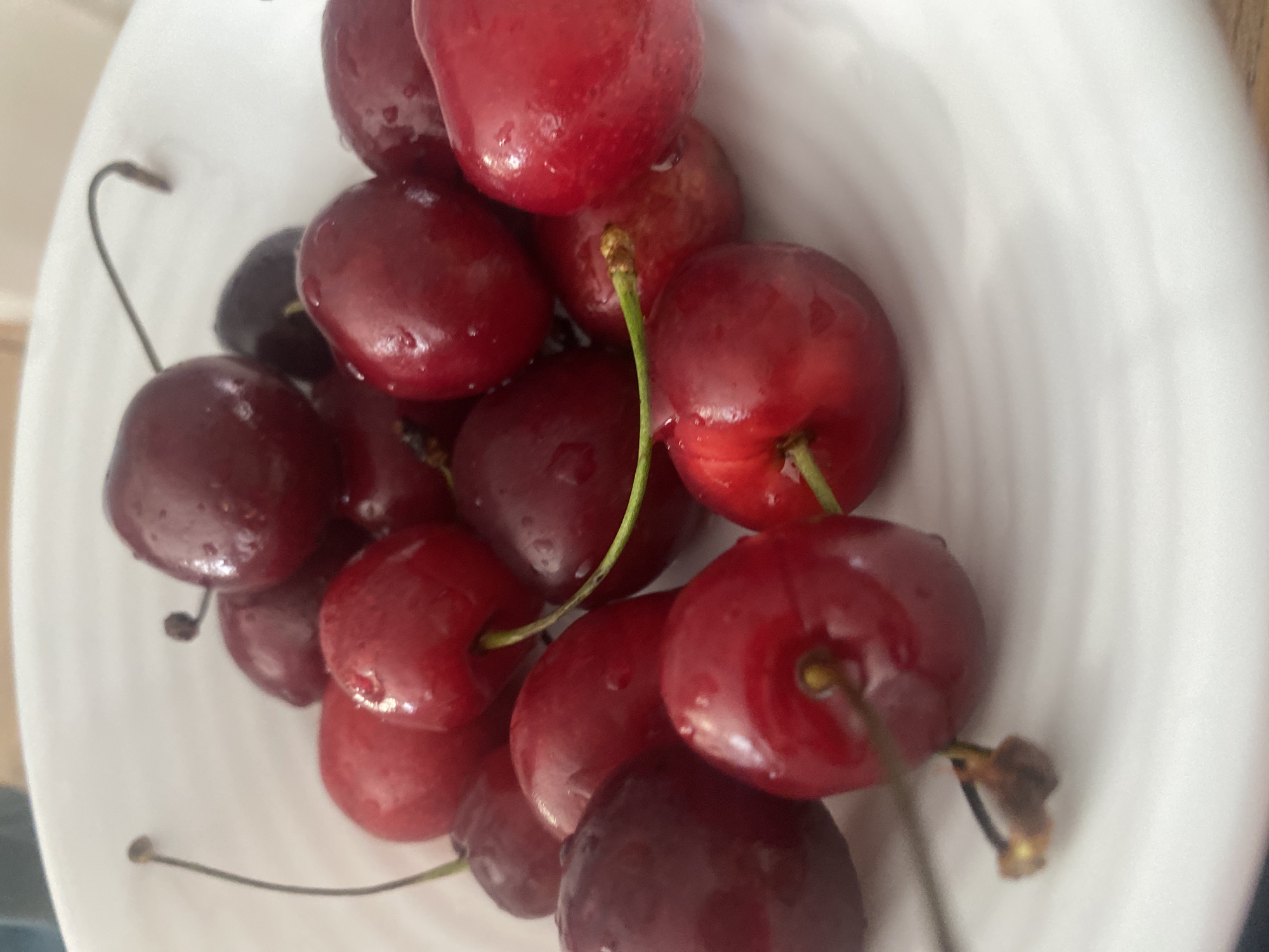 A close-up shot shows a white bowl filled with dark red cherries. The cherries are round and plump, with some appearing slightly wet, suggesting they might have been washed. A few have thin stems attached, curved in various directions. The bowl has a ridged interior texture visible in the background. The lighting is soft, illuminating the glossy surfaces of the cherries and casting subtle shadows. The composition is focused on the fruit, with no other objects or people visible. The scene appears to be an indoor setting, possibly a kitchen or dining area. The overall impression is one of freshness and simple natural beauty. There is no visible text in the image.