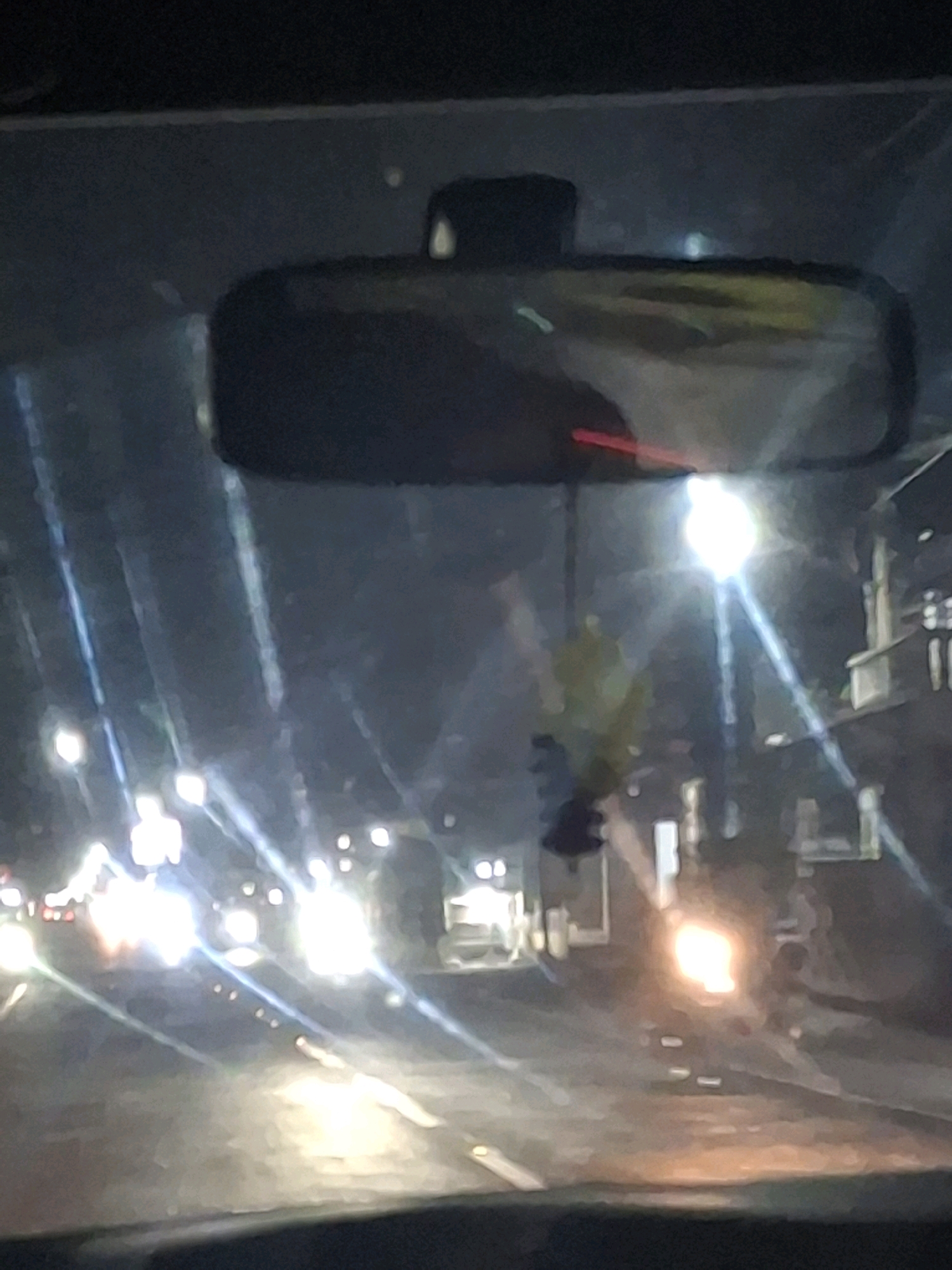 The image depicts a view from inside a vehicle at night, likely on a street in Lumajang, Indonesia. The primary focal point is the rearview mirror, reflecting a blurry, indistinct image of the road ahead. Outside the vehicle, streetlights and vehicle headlights illuminate the dark environment, creating streaks of light and lens flares across the windshield. Buildings are visible on the right side of the frame, appearing as blurred structures in the background. A blurry object, possibly an air freshener, hangs from the rearview mirror. The overall scene is characterized by low light conditions, motion blur, and reflections, suggesting the vehicle is in motion on a city street.