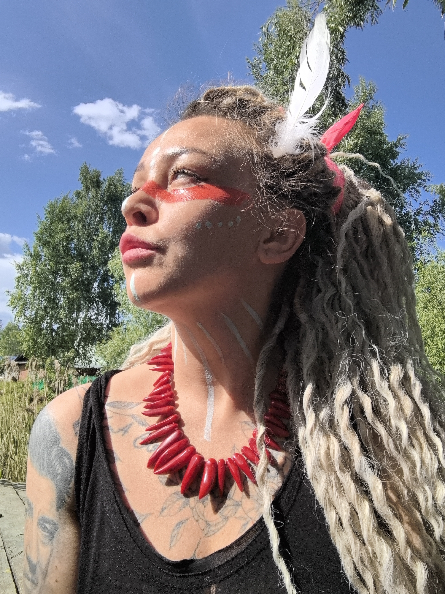 A woman with dreadlocks and face paint is looking up at the sky. She has red paint across her forehead and cheeks, white dots on her cheekbones, and white stripes on her neck. She wears a red necklace and a black tank top. Her left shoulder is tattooed. She has a white feather and a red piece of fabric in her hair. In the background are trees and a blue sky with clouds. The location is Novyi, Russia.