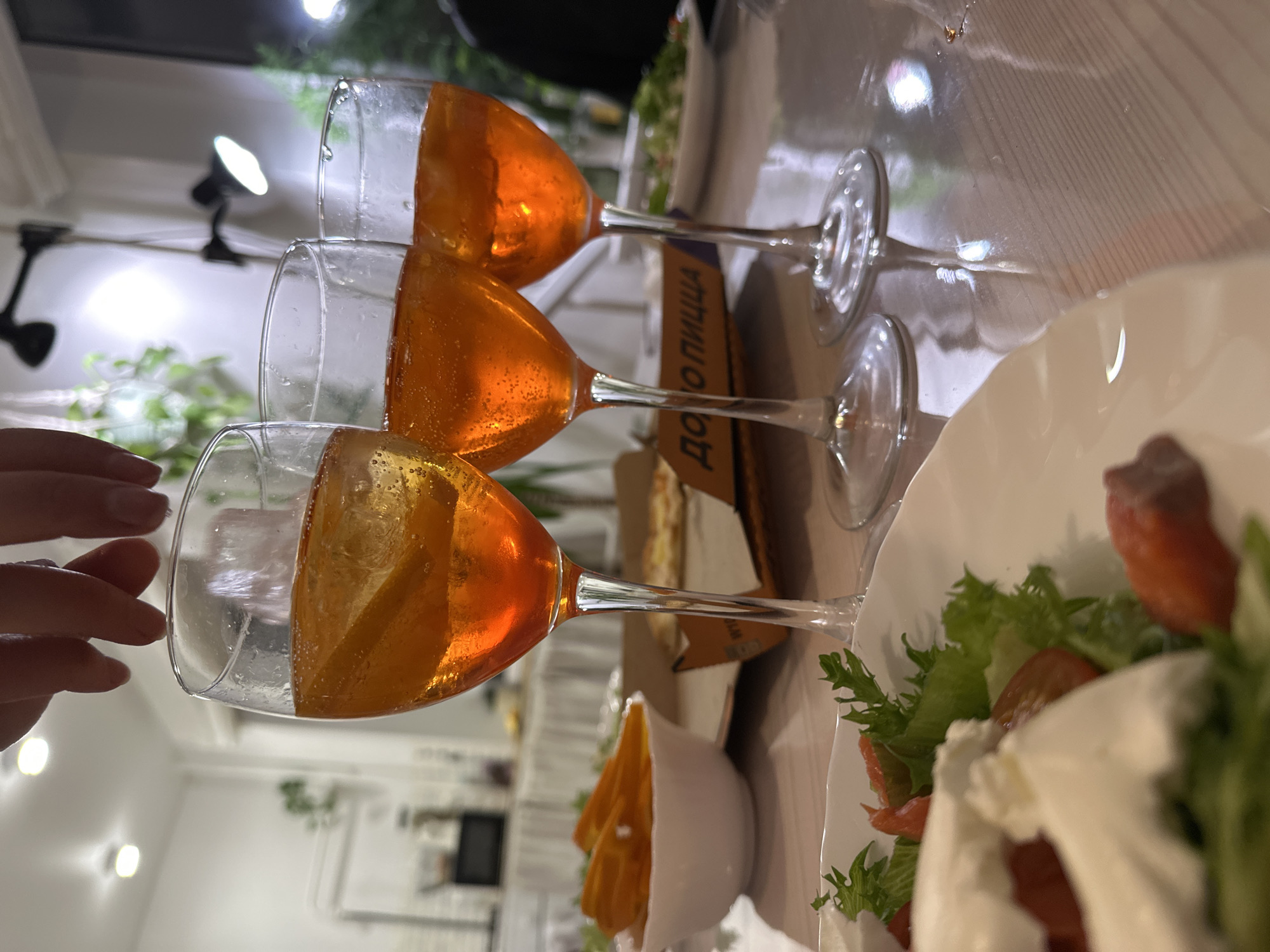 The image depicts a table setting with three glasses of Aperol Spritz, a white bowl of orange slices, a plate of salad with what appears to be mozzarella cheese and salmon, and a pizza box. The location is identified by text on the pizza box as being in Chaikovsky, Russia ("до пицца"). A hand with light skin and manicured nails is visible in the lower left corner. The table surface is light-colored wood. The background shows interior lighting fixtures and a glimpse of greenery and a television. The perspective suggests the image was taken close to the table.