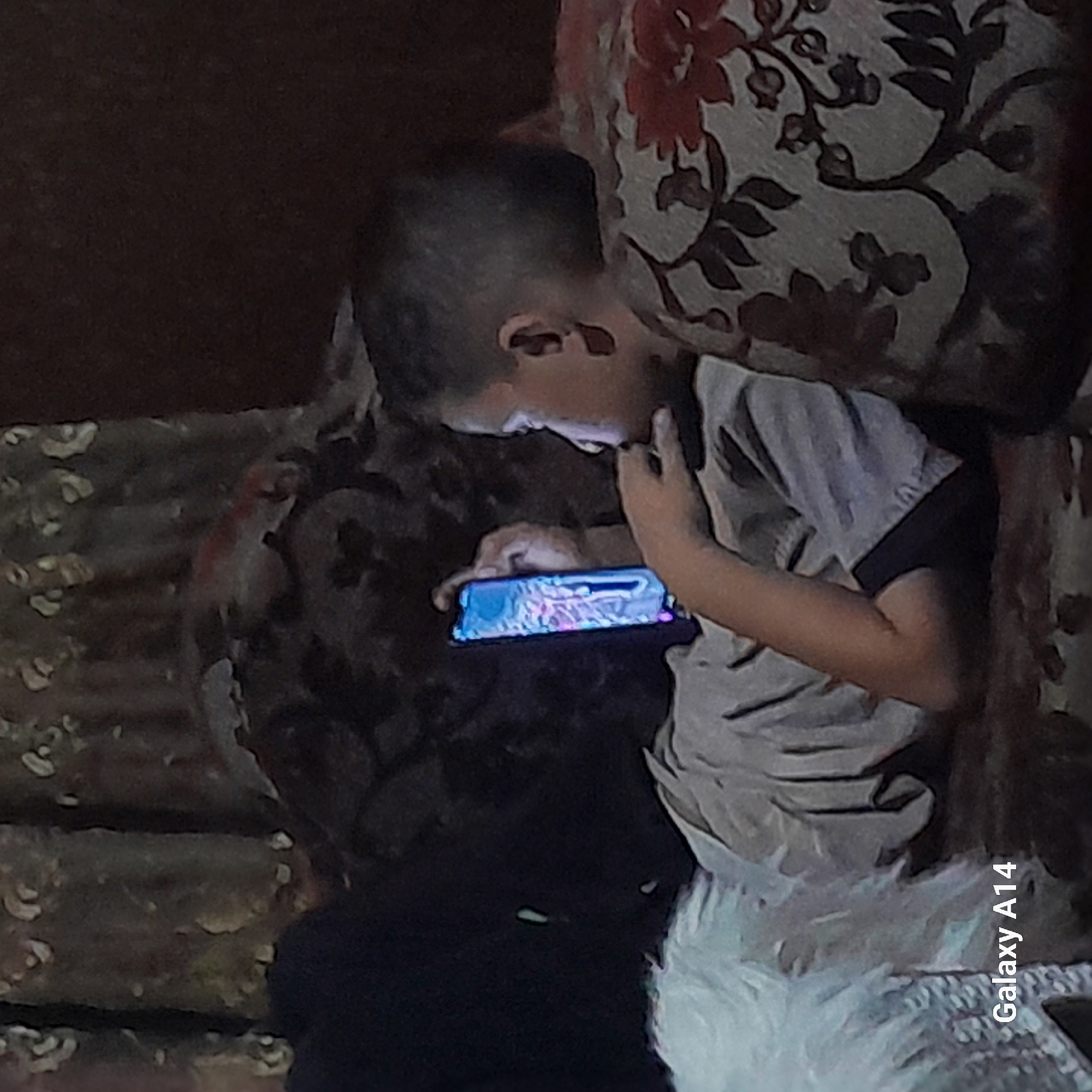 A young boy is lying on a couch, looking at a smartphone. He is wearing a grey t-shirt and dark shorts. The couch is covered with a patterned fabric. A fuzzy white blanket or cushion is visible to the left of the boy. The room appears to be dimly lit. The text "Galaxy A14" is overlaid on the bottom left of the image. This image was taken in Khan Yunis, Palestinian Territories.