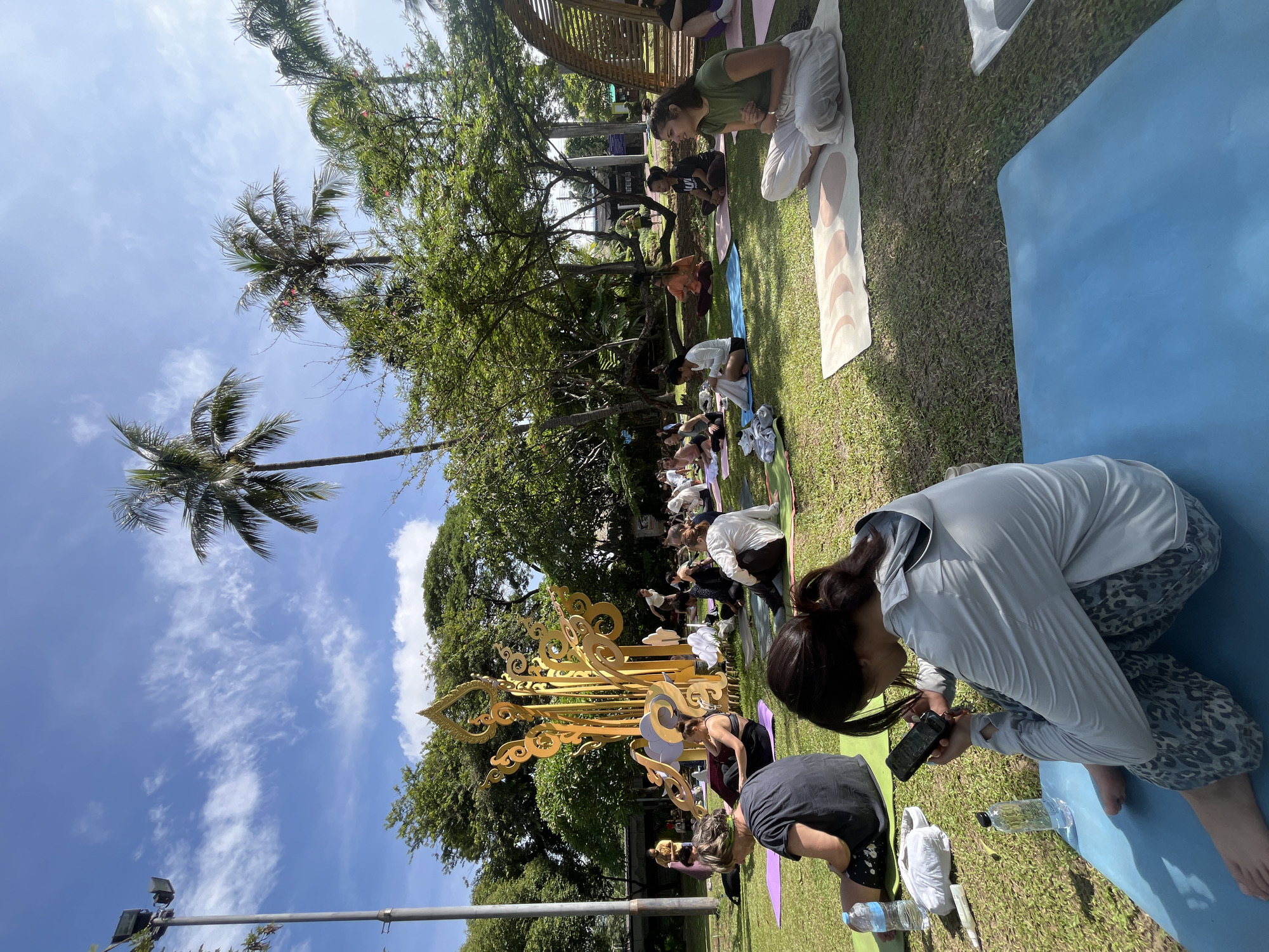 A group of people are sitting on yoga mats in a park on a sunny day. A woman in the foreground is looking at her phone. In the background, there is a large, ornate golden sculpture. Palm trees and lush green foliage are visible, suggesting a tropical or subtropical environment. The scene appears to be a yoga or meditation session taking place outdoors. The presence of palm trees and a large, decorative golden sculpture is consistent with the aesthetics found in Chiang Mai City Municipality, Thailand, which often features lush greenery and intricate artistic installations in its public spaces and parks.