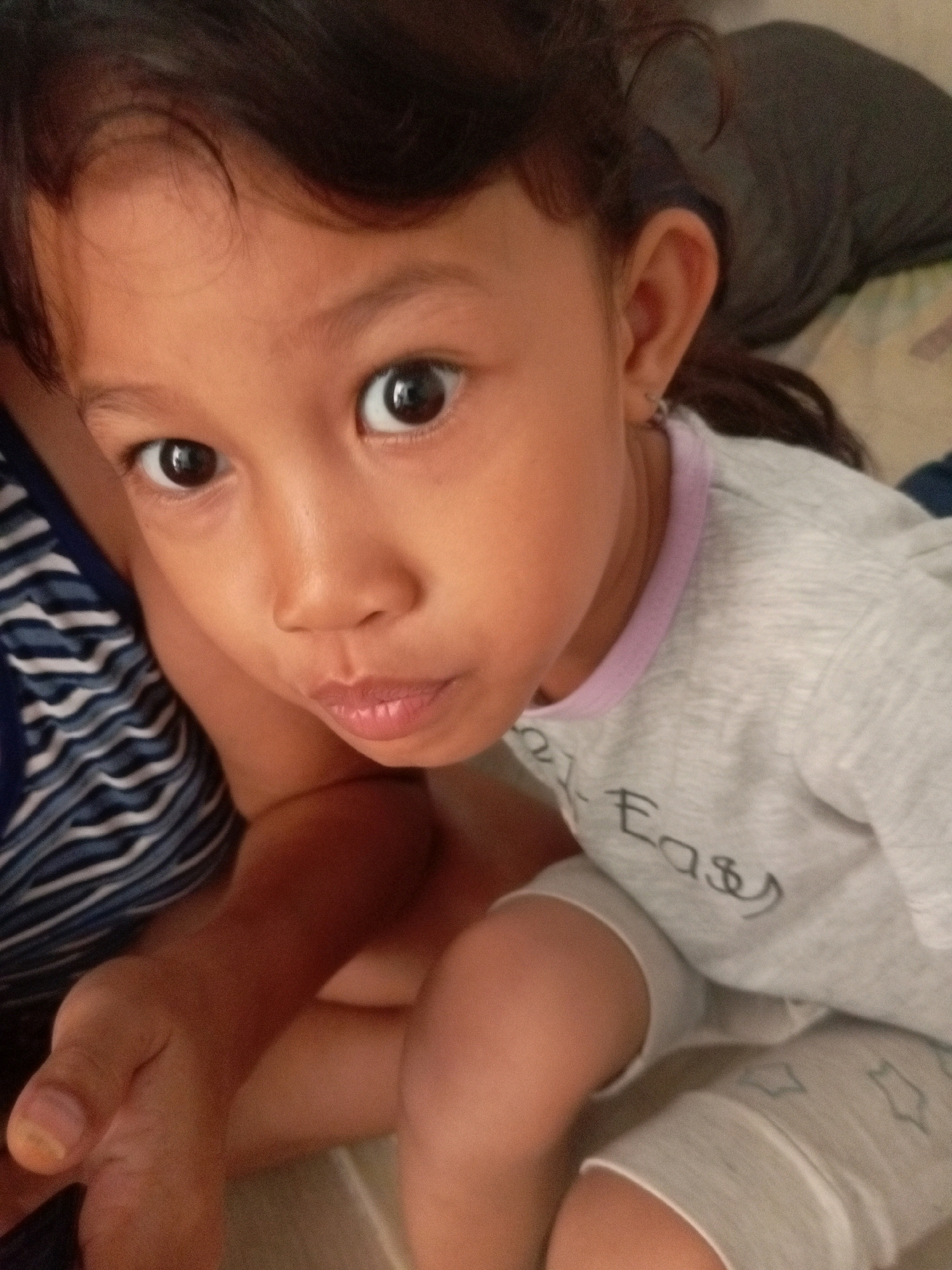 This is a close-up, low-angle medium shot of a young East Asian girl, likely a toddler or preschooler, indoors. She has dark hair and fair skin. Her face is the primary focus, with large, dark eyes looking directly at the camera, and her lips pursed. She is wearing a light grey t-shirt with a pink collar, and light grey shorts that have small, outlined star shapes printed on them. A portion of a blue and white striped garment is visible on the left side of the frame, suggesting she might be wearing it or it belongs to someone else nearby. A hand, presumably belonging to an adult, is visible in the lower left corner, resting on her leg. The background is blurred, showing a darker fabric, possibly a blanket or clothing, and a lighter surface that could be a bed or couch. The lighting is soft, suggesting an indoor setting during daylight hours. There is visible text on the girl's t-shirt, partially obscured, but the word "Easy" is discernible. The overall impression is one of a candid, intimate portrait.
