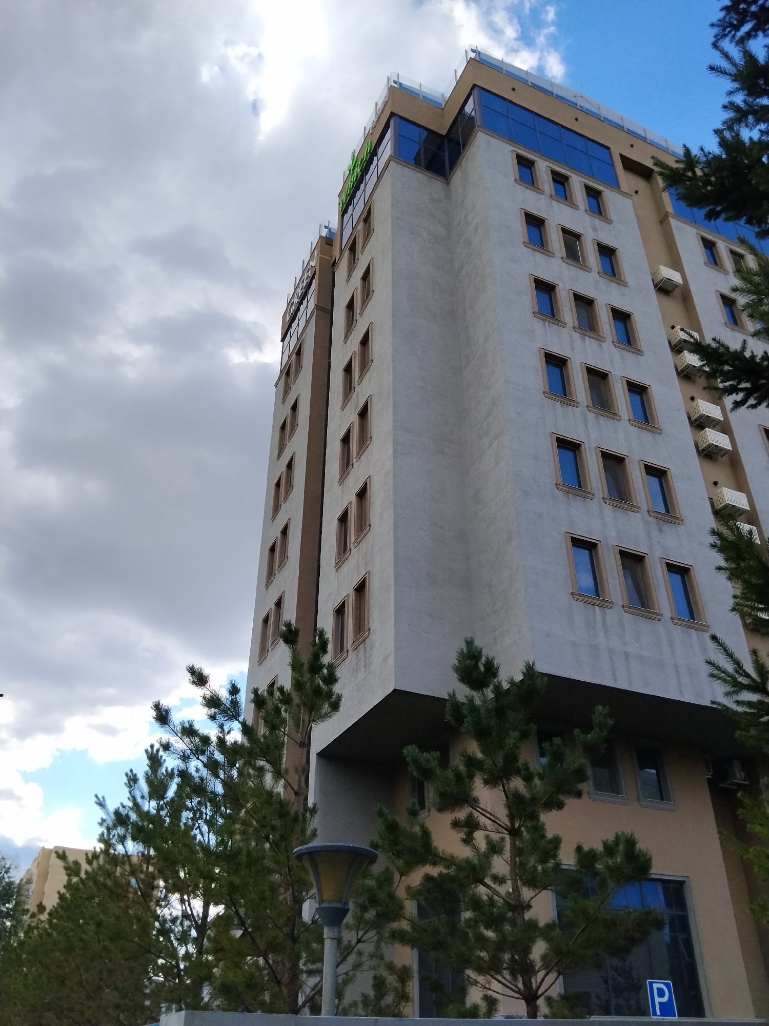The image shows a tall, modern building with a light-colored facade and brown accents. The building has many windows, some of which are tinted blue. The top of the building features a glass facade and a railing. Green trees with pine needles are in the foreground, partially obscuring the lower floors of the building. The sky is cloudy, with patches of blue sky visible. A street lamp is visible in the lower right corner. A blue sign with a white "P" symbol is also visible near the bottom right. This building is the Park Inn by Radisson Kokshetau, located in Kokshetau, Kazakhstan.