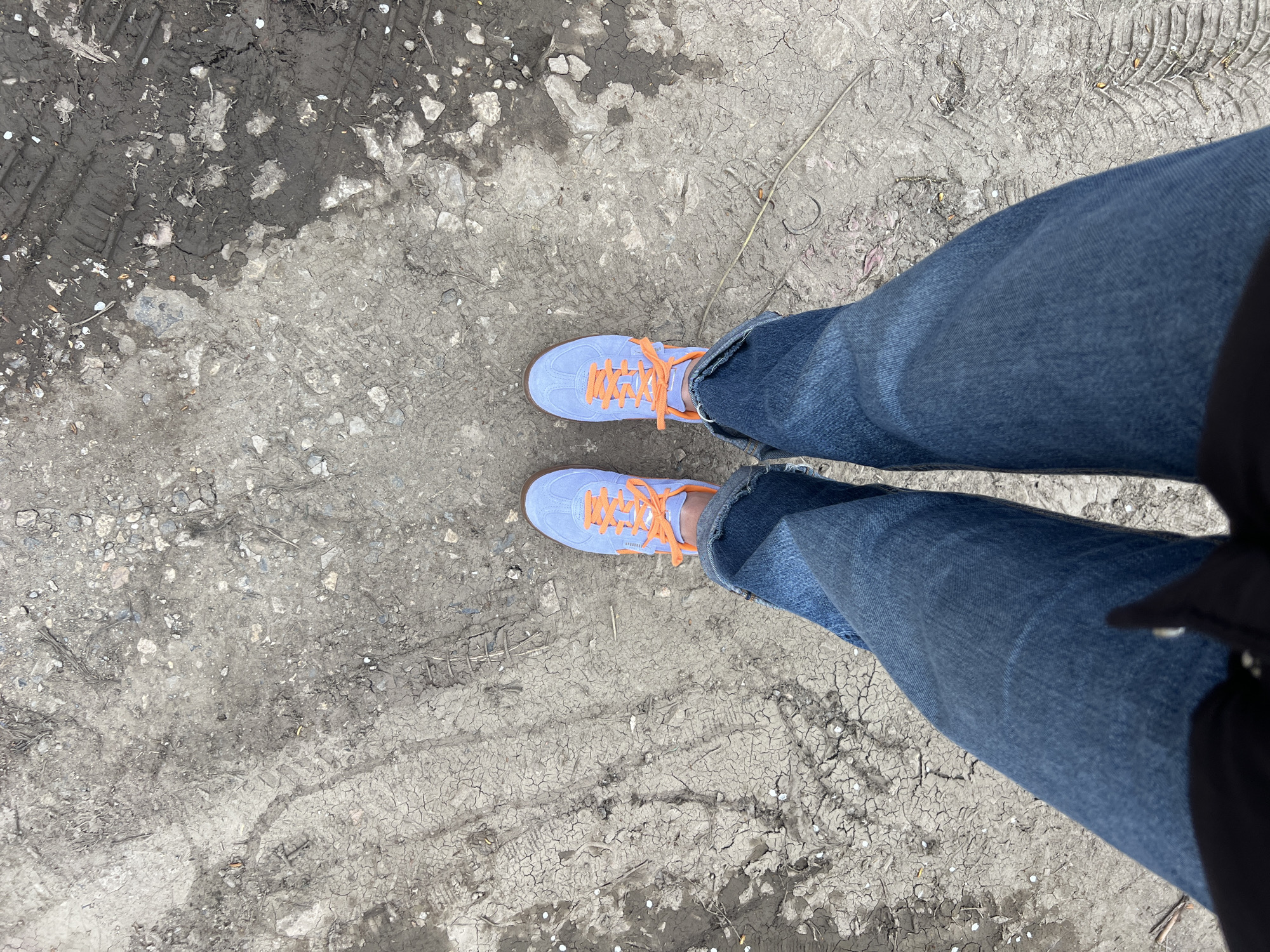 The image shows a top-down view of a person standing on cracked, dirty ground. The person is wearing blue jeans and light blue sneakers with orange laces. The ground appears to be a mixture of dry dirt and mud with tire tracks visible in the upper left corner. The image was likely taken outdoors in an unpaved area. The location in the city of Sofia, Bulgaria cannot be determined from the information within this image.