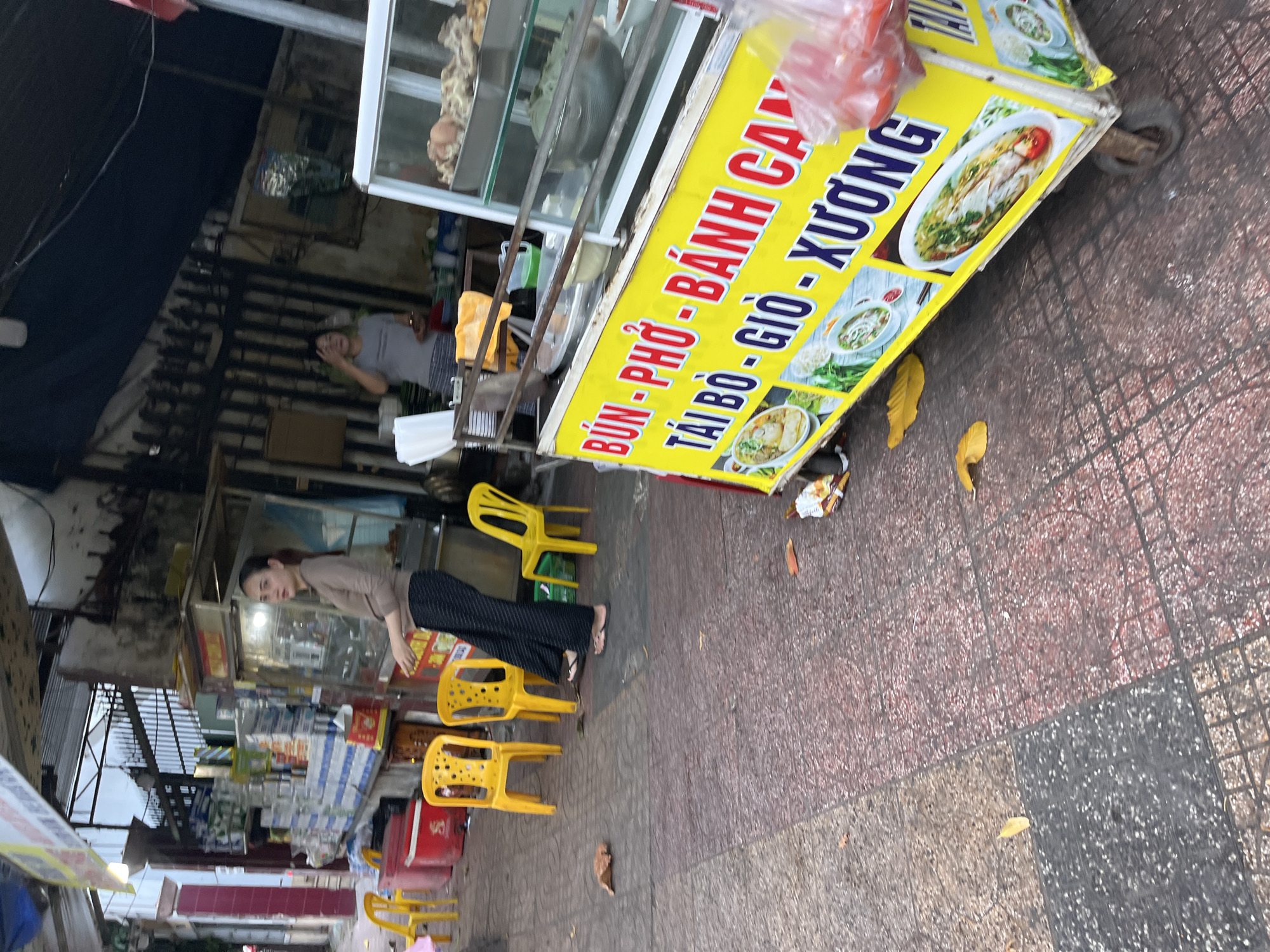 In Nha Trang Ward, Vietnam, a street vendor's cart with a bright yellow sign is positioned on a patterned tiled sidewalk. The sign displays Vietnamese text and images of noodle soup dishes, indicating the sale of food items such as "BUN - PHO - BANH CAN". A woman wearing a light brown long-sleeved shirt and dark patterned pants stands near the cart, looking towards the left of the frame. In the background, a glass display case on the cart holds various food items, and a person is visible behind it, appearing to be preparing or serving food. Several yellow plastic chairs are placed on the sidewalk, suggesting an outdoor seating area for customers. The environment appears to be a bustling street scene with structures and other vendors visible in the distance.
