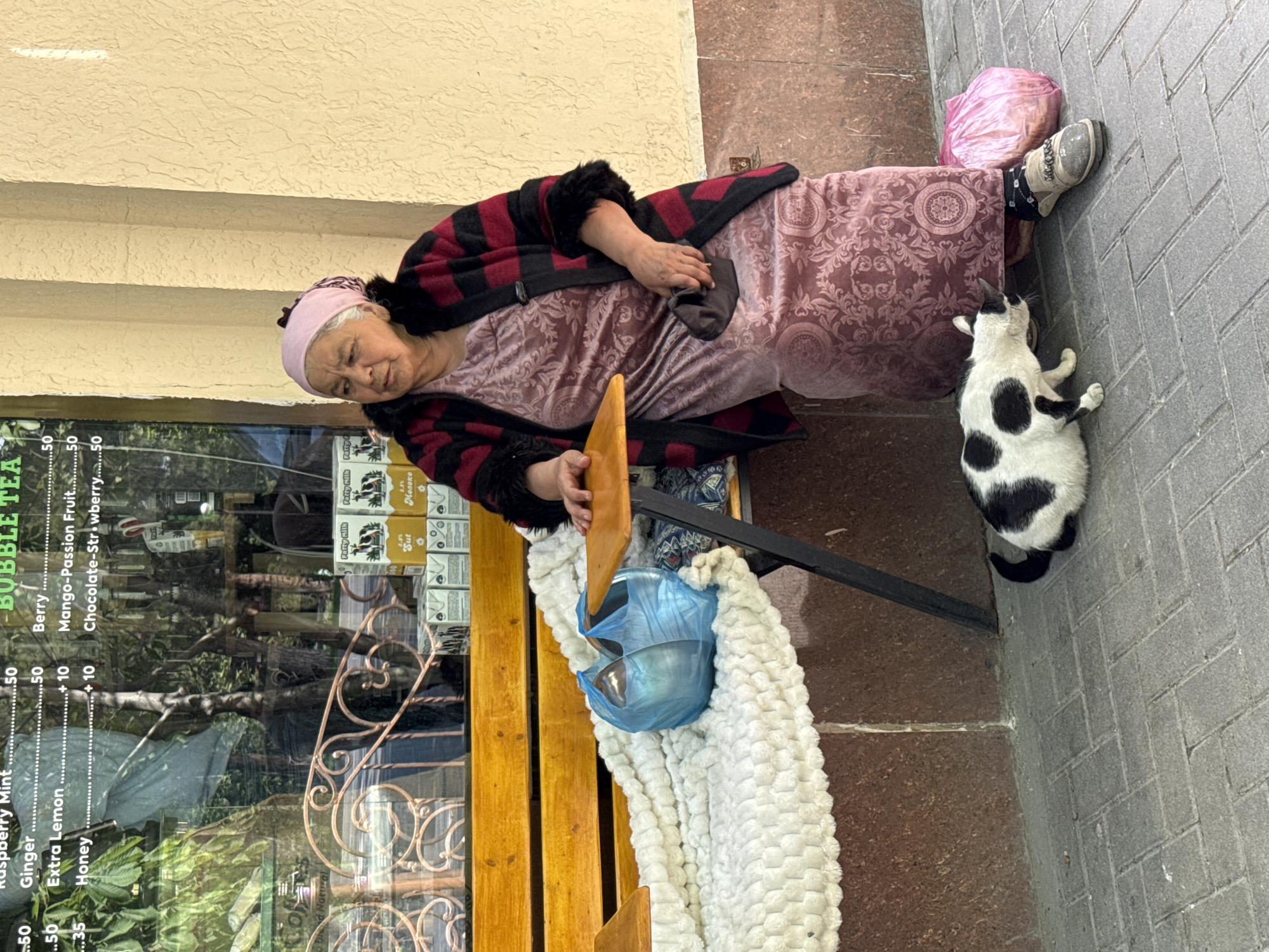 The image depicts an elderly woman sitting on a wooden bench outside a shop in Samarkand City, Uzbekistan. She wears a pink headscarf, a floral patterned dress, and a red and black striped jacket. On the bench, next to the woman, there's a blue plastic bag and a white blanket. A black and white cat stands on the grey brick sidewalk next to the bench. The shop window displays a menu for "Bubble Tea," listing flavors like Raspberry Mint, Ginger, and Mango-Passion Fruit.