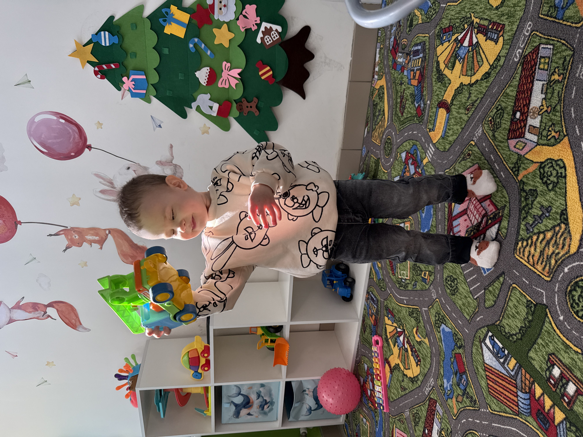 A young child in Rusanovo, Russia, wearing a light-colored sweater with black and white cartoon animal designs and dark-colored pants, is playing with a colorful toy car/truck with a green base and yellow and blue car parts on top. The child is standing near a white shelf unit filled with various toys. Behind the child is a wall decorated with fox and balloon decals, as well as a felt Christmas tree adorned with felt decorations. A playmat depicting a city scene with roads and buildings is on the floor.
