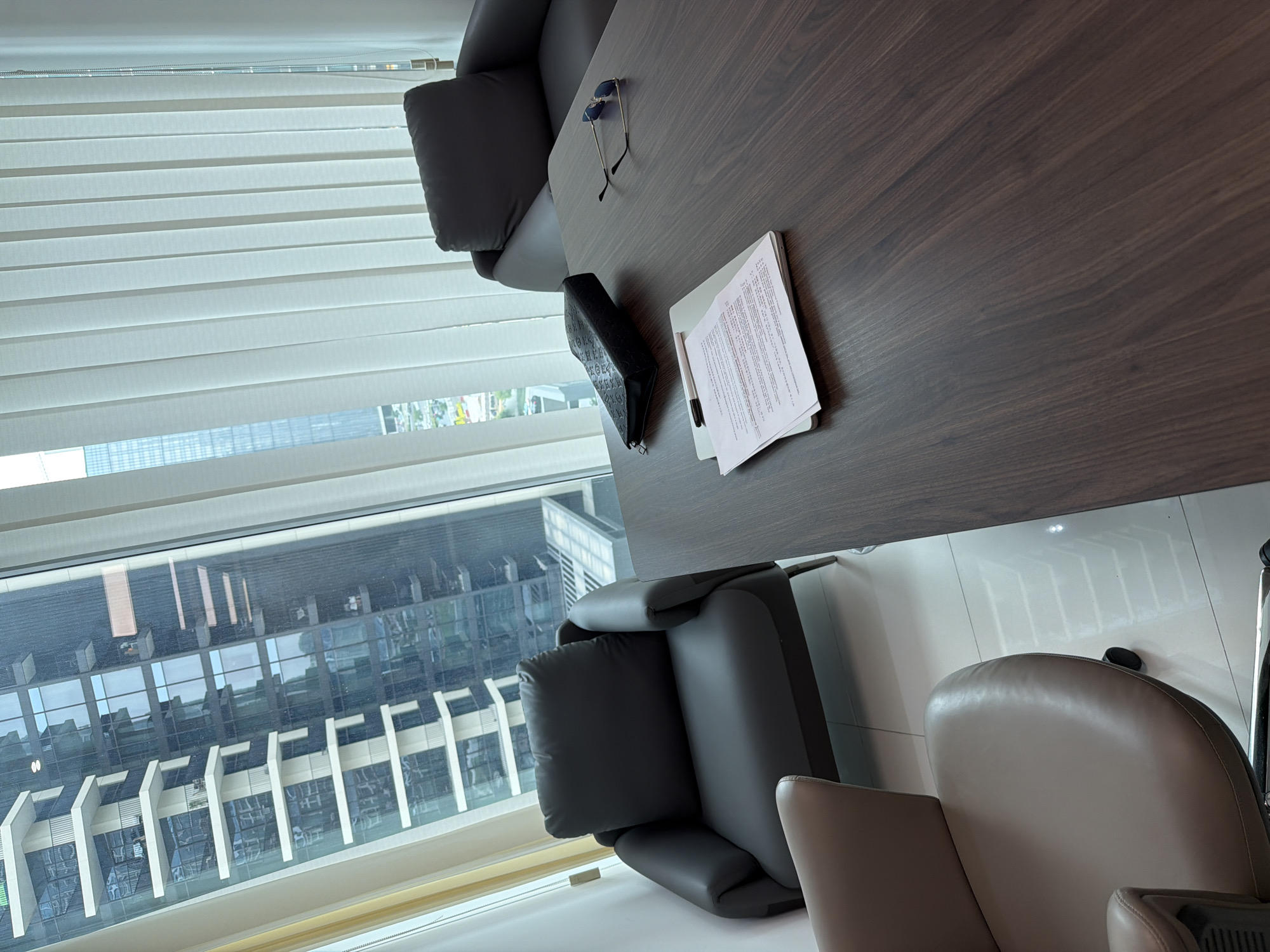 A high-angle, indoor shot depicts a portion of a meeting room in Business Bay, United Arab Emirates. Visible through a large window are numerous high-rise buildings, characteristic of the urban landscape. The window is partially covered with horizontal blinds.
A dark-brown wooden table dominates the right side of the frame. On the table are several items: a pair of sunglasses, a black wallet, a pen, and a stack of papers.
Two dark-gray leather chairs with cushions sit near the table. A light-beige leather office chair is partially visible at the bottom-right. No people are visible. The floor appears to be light-colored tile.