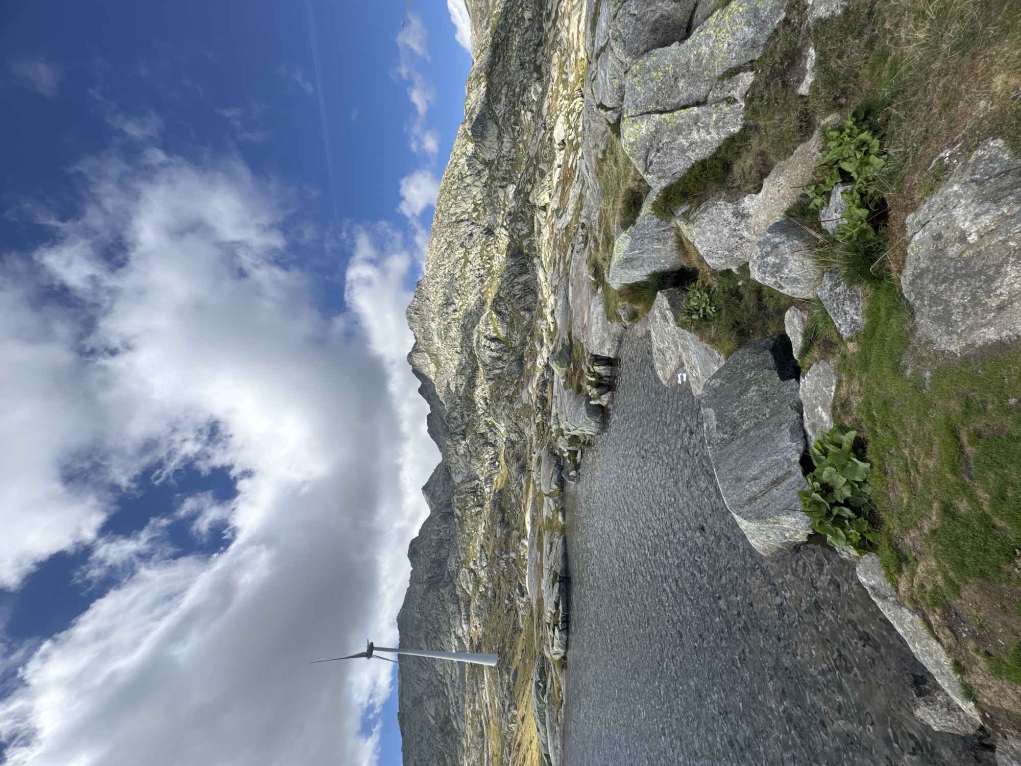 The image captures a serene mountain landscape in Airolo, Switzerland. A body of dark, rippling water occupies the left foreground, with the shore made up of large, grey rocks and patches of green grass. In the mid-ground, a white wind turbine stands tall on a rocky, barren slope. Behind the turbine, rugged, grey mountains rise sharply, their peaks partly obscured by clouds. The sky above is a mix of bright blue with scattered white and grey clouds, suggesting a dynamic weather system. The overall impression is one of a remote, natural environment with a prominent man-made structure integrated into the landscape. There are no people visible in the image.