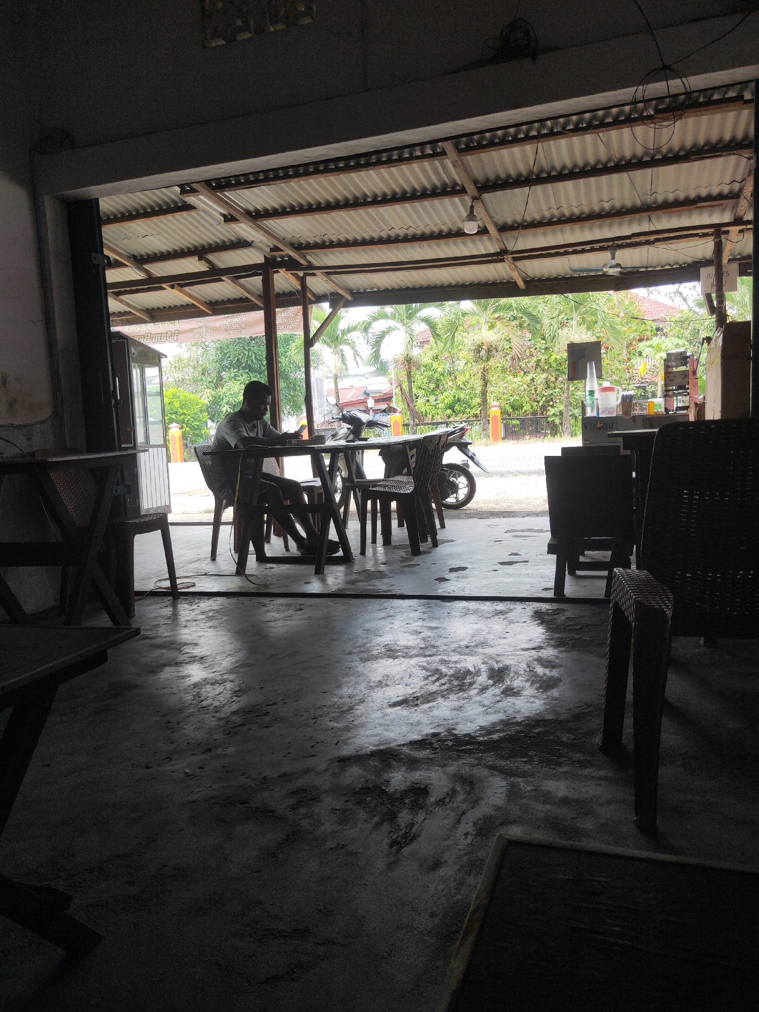An outdoor cafe area is depicted with a man sitting at a table and looking at his phone. The cafe appears to be in Sintang, Indonesia, as indicated by the presence of lush tropical vegetation, palm trees, and architecture consistent with the region. The cafe is open-air, with a corrugated metal roof supported by wooden beams. Wicker chairs and tables are arranged throughout the space, and a motorcycle is parked nearby. The floor is concrete and appears wet in places, reflecting the ambient light. The overall impression is one of a casual, slightly weathered, outdoor dining establishment in a warm, humid climate.