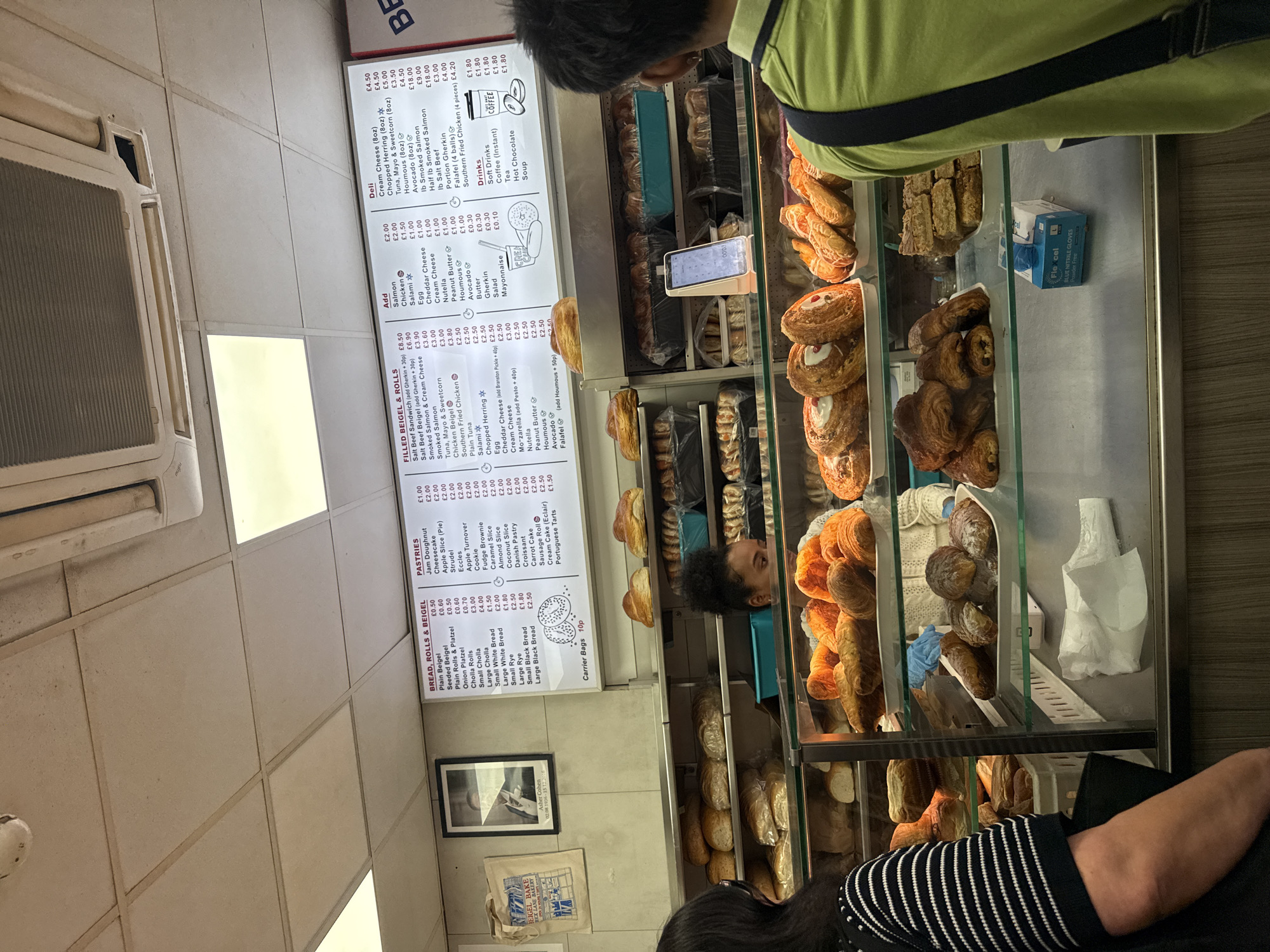The image shows a customer ordering food at Beigel Bake, a bakery on Brick Lane in London, United Kingdom. The counter is filled with pastries, croissants, and other baked goods. A menu board is visible, listing various beigels, rolls, and pastries, along with their prices. The customers are looking at the food on display, and a staff member is present behind the counter. Packaging for gloves can also be seen on the counter.