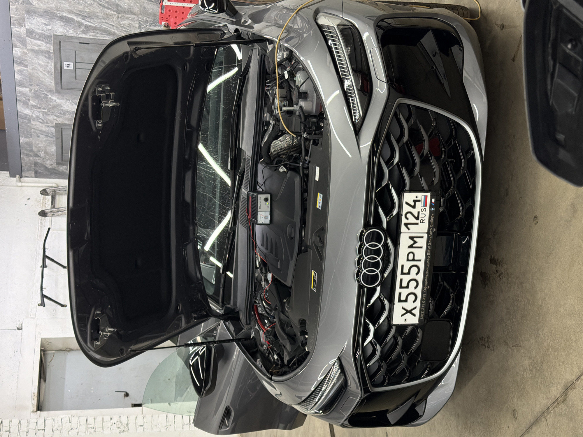 The image shows a gray Audi car with its hood open, revealing the engine compartment. A battery charger is connected to the engine. The car has a Russian license plate that reads "X555PM 124 RUS". The license plate also contains the phone number (383) 319 0 319 and mentions "Официальный дилер BMW ЭлитаАвто Сибирь", indicating a BMW dealership. The background is a garage with gray walls. The location is likely Krasnoyarsk, Russia, given that the area code for Krasnoyarsk is 124 and the dealership mentions Siberia.
