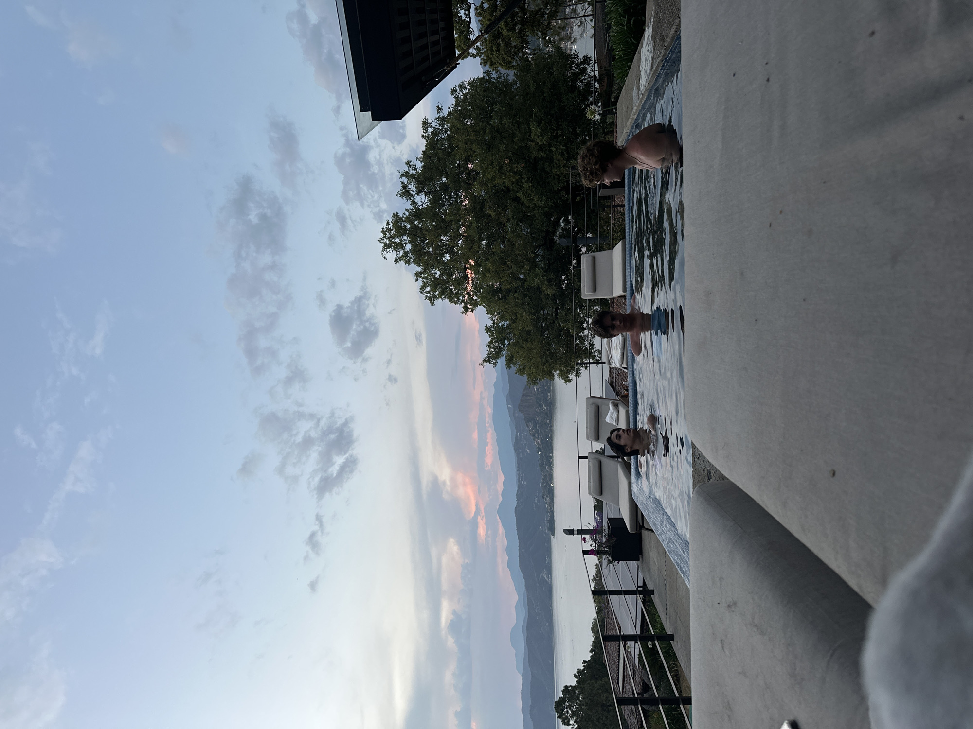 The image shows a view from a high vantage point, potentially a balcony or terrace, overlooking a pool and a distant landscape. In the pool, several people are visible, suggesting a leisurely activity. The backdrop includes a body of water, mountains, and a sky with clouds and hints of sunset colors. The scene appears to be located in a setting with a combination of natural and architectural elements. The exact city or specific location is unknown based on the image alone.
