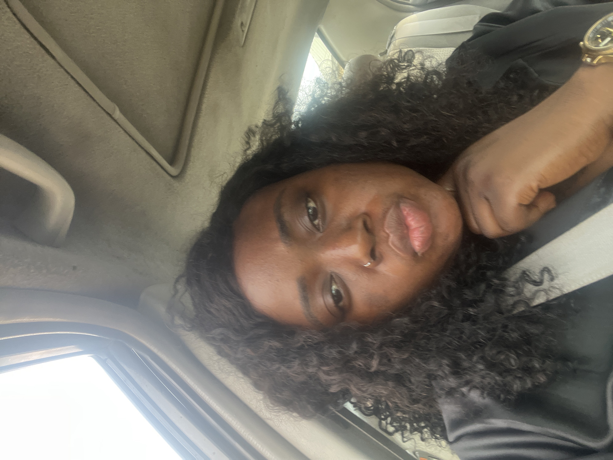 A woman with dark curly hair and a nose piercing is seated inside a car. She is wearing a black top and a gold watch. The car's interior is beige. The location is not discernible as Abuja, Nigeria.