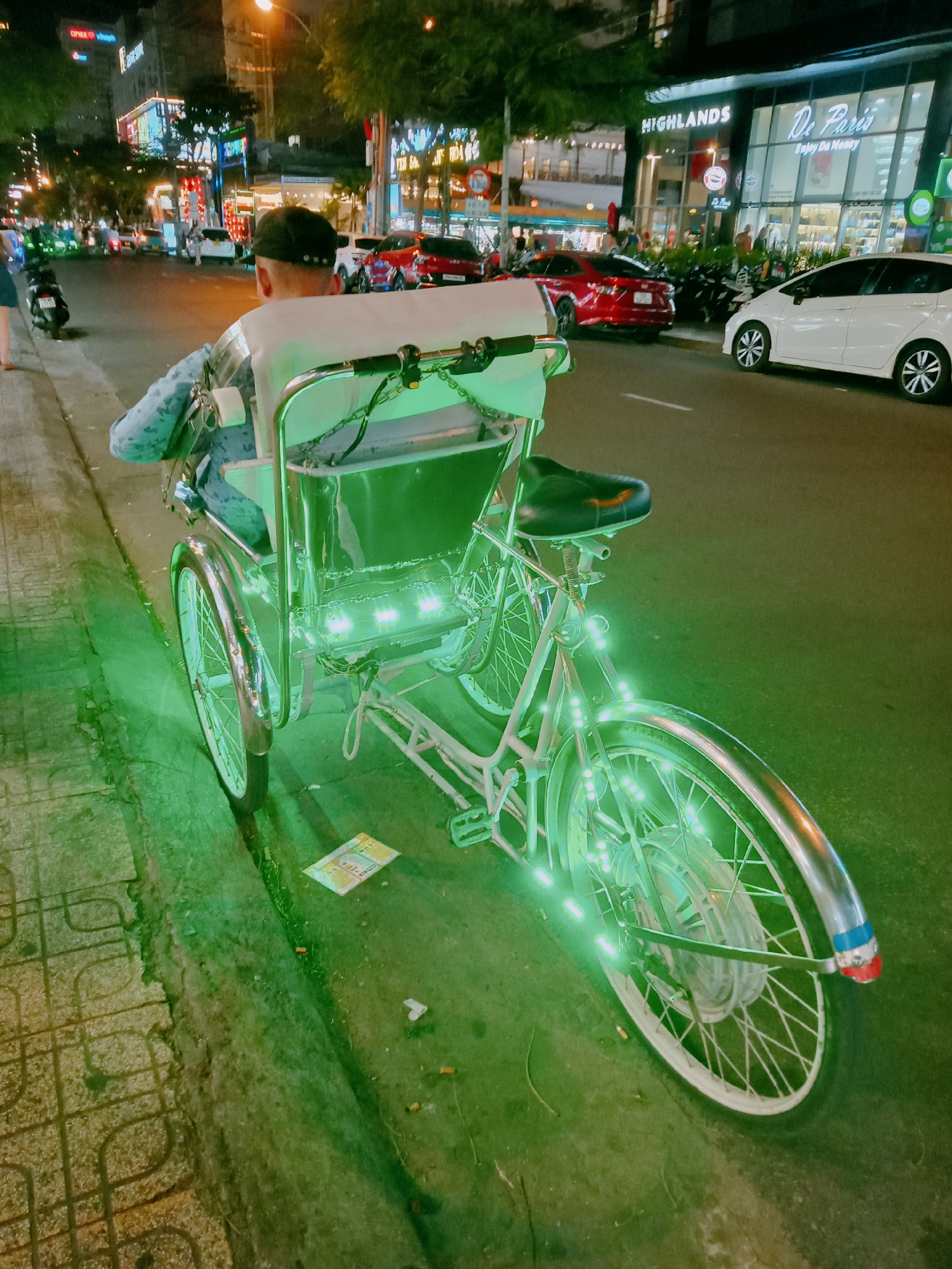 An illuminated cyclo sits parked on a sidewalk in Nha Trang Ward, Vietnam. The cyclo, a three-wheeled bicycle taxi, is lit with green lights and faces away from the viewer. The driver, wearing a black baseball cap and a light blue patterned shirt, is seated. The cyclo's seat is black. A piece of paper lies on the ground nearby. In the background, the street is illuminated by streetlights and buildings. A white car and a red car are visible on the road, as well as motorbikes. Buildings with lighted signs for "HIGHLANDS" and "De Paris" can be seen across the street.
