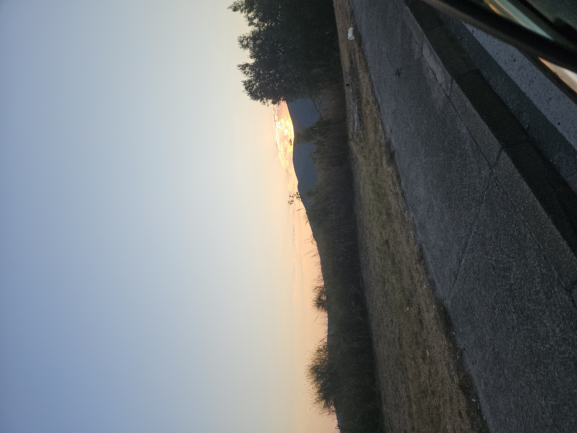 A wide shot captures a tranquil scene of a sunset over a mountain range. The sky is a gradient of pale blue to soft orange, with the sun partially obscured by clouds, casting a warm glow. In the foreground, a dry, grassy field slopes gently towards a paved road with a curb. Dark green trees and shrubs dot the landscape, creating a natural border between the field and the mountains. The overall impression is one of peaceful solitude, with no people or animals visible. The image appears to be taken from a moving vehicle, as indicated by the blurred edge of a car window in the lower right corner.