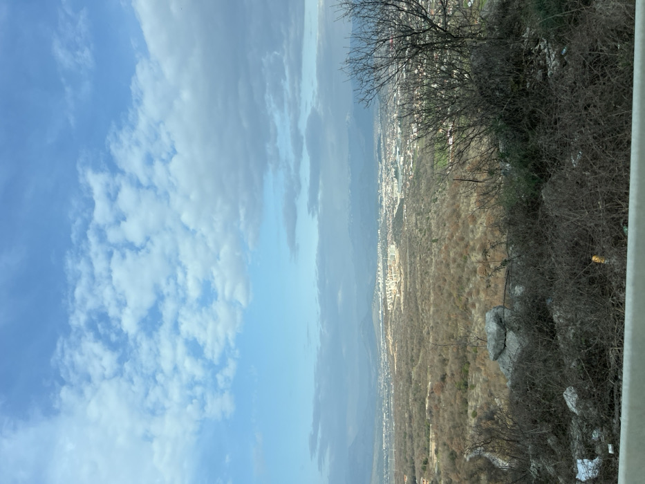 This image presents a panoramic landscape view from an elevated position overlooking the city of Gornji Kokoti, Montenegro. The immediate foreground features a white horizontal barrier, likely a guardrail, spanning the bottom of the frame. Above this barrier, the terrain is covered with dense, dry, dormant shrubbery and scattered detritus, including two prominent grey rocks centrally located and a leafless deciduous tree with dark branches on the right. Visible among the sparse vegetation are small pieces of white plastic and a yellow cylindrical object.
The midground consists of an extensive, undulating landscape dominated by numerous bare deciduous trees and shrubs, displaying a muted palette of browns and tans, indicative of winter or late autumn. A faint winding road or path is visible descending through this terrain on the far left.
In the background, the city of Gornji Kokoti sprawls across a broad plain, characterized by a concentration of urban buildings and structures. Further in the distance, mountains, some with visible snow patches on their peaks, define the horizon line, particularly towards the right side of the frame. Atmospheric haze partially obscures the most distant geographical features.
The sky occupies the upper portion of the image, displaying a blend of clear blue and various cloud formations. White cumulus clouds are scattered across the sky, with a more substantial band of grey clouds extending horizontally across the middle-right. The scene is illuminated by natural daylight. No individuals, specific actions, or interactions are discernible.