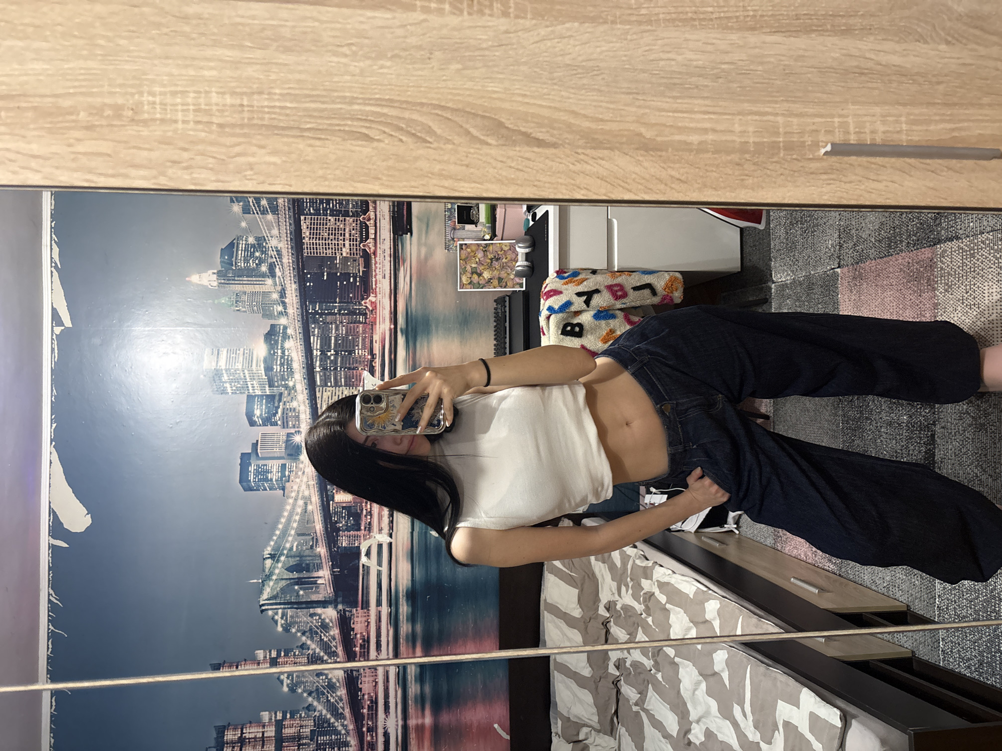 A young person stands in front of a mirror, taking a selfie. The person is wearing a white cropped tank top and dark denim jeans. Their dark hair is long and straight. The phone is visible in the person's hand, obscuring part of their face.
The reflection in the mirror shows a bedroom setting. Behind the person is a wallpaper depicting a cityscape. A bed with a grey and white patterned duvet is visible below. To the right of the bed is a set of drawers. Other items are visible in the background, including a colorful cushion and various objects on a shelf.
The location is likely indoors in a residential setting, but there is no direct evidence that this is in the city of Sofia, Bulgaria.