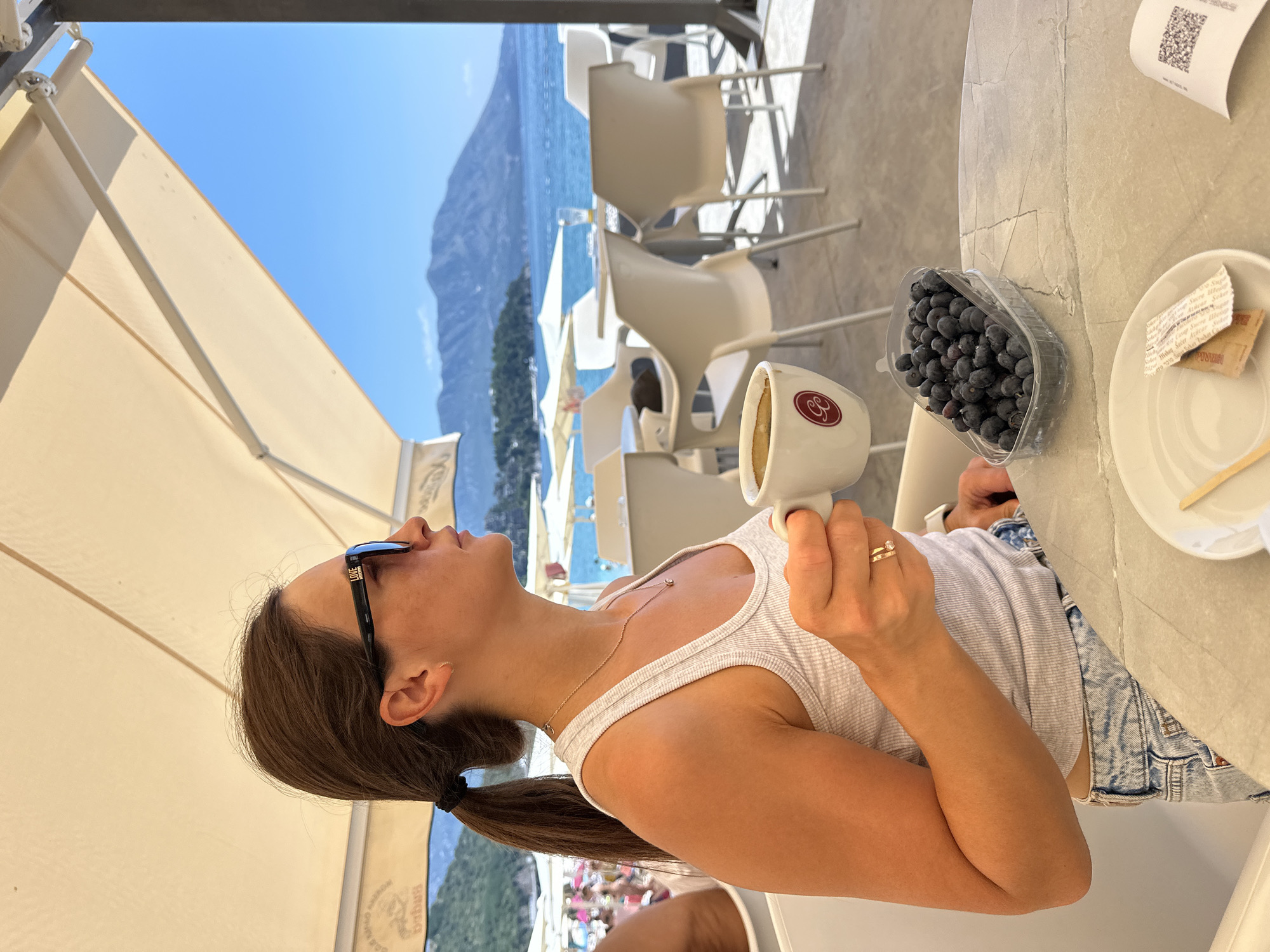A woman with brown hair pulled into a ponytail, wearing a white tank top, denim shorts, and sunglasses, sits at a table outdoors, holding a white cup of coffee and looking out at the sea. A plastic container of blueberries is on the table next to her, along with a small plate with sugar packets and a stir stick. The background shows a beach with umbrellas and people, and in the distance, a body of water and mountains. The scene appears to be in Budva, Montenegro, likely at a beachfront cafe.