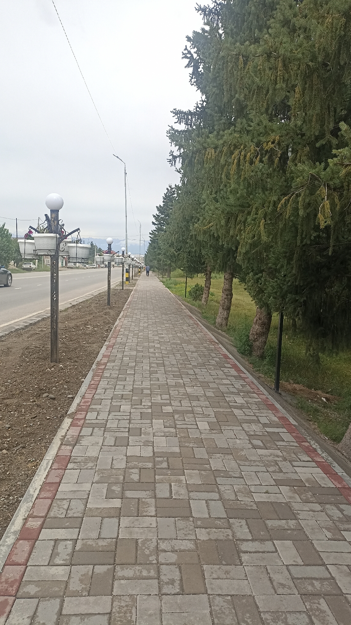 A paved pedestrian walkway, bordered by trees on the right and a road with sparse vehicular traffic on the left, stretches into the distance under an overcast sky. The walkway is constructed of interlocking grey and red pavers, laid in a herringbone pattern. Along the left side of the walkway, a row of decorative lampposts with globe-shaped lights are spaced out. Beyond the lampposts is a paved road with at least one car visible. To the right of the walkway, a line of mature evergreen trees with thick trunks and dense foliage forms a natural barrier. The ground beyond the trees is a grassy area. In the distance, a single person is walking along the pedestrian path. The overall scene depicts an urban or semi-urban environment, likely a streetscape or park area in Ak-Suu, Kyrgyzstan.