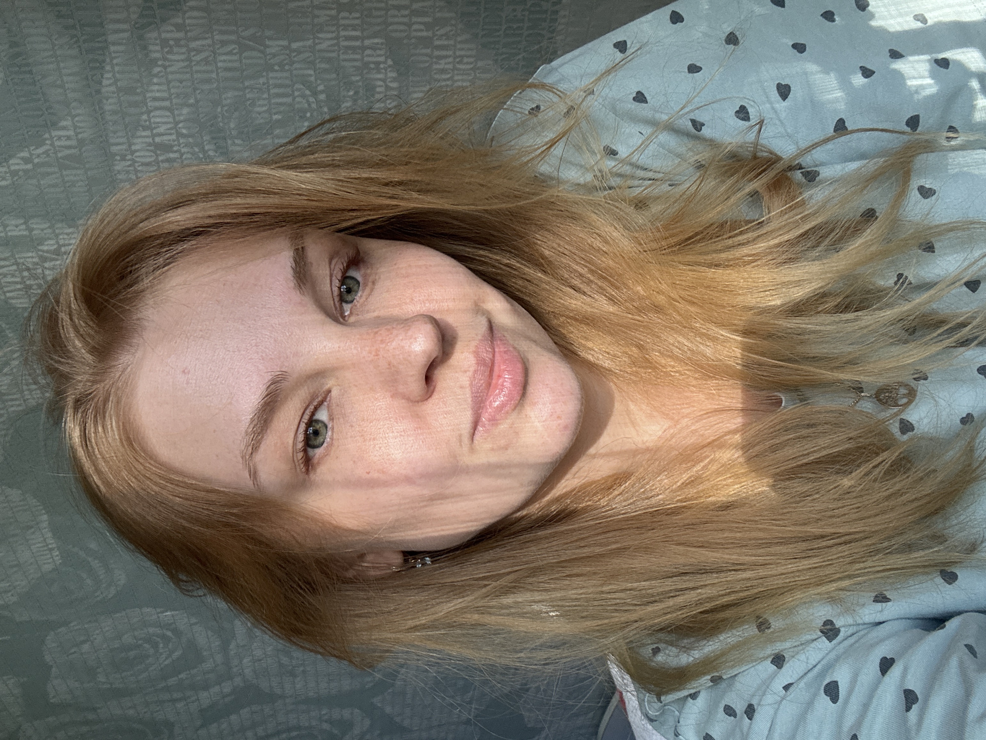 A young woman with long, light blonde hair, green eyes, and freckles is depicted in a close-up, high-angle shot, appearing to lie down. She has a relaxed expression with a gentle, closed-mouth smile. Strong, directional sunlight illuminates her face and hair, casting distinct linear shadows across her features, indicating a bright daytime setting, likely morning or late afternoon.
She is wearing or is covered by light blue fabric patterned with small dark heart shapes, suggesting sleepwear or bedding. Her hair is spread out around her head, catching the sunlight. Behind her head and to the left, another fabric, possibly a pillowcase or headboard, features a decorative pattern incorporating fragmented and jumbled text. Visible words and phrases from this pattern include "UGH/YELL", "LAND NOR", "ENARECKA", "A COMPACT", "BY AND THES MANG", "BALOWERING PLA", "BETASPECIESANDT", "SWITHST", "ABGEAND/SHO", "NA WITH:SM", "BADSAREN", "ZINGMA", "EMETER", "WIDE RANGE", "SCORE", "UP OF PLA", "CKLES.FF", "JUGHYAT", "OLAND", and "ENARE". The scene suggests a tranquil, indoor moment, likely in a bedroom in Rusanovo, Russia.