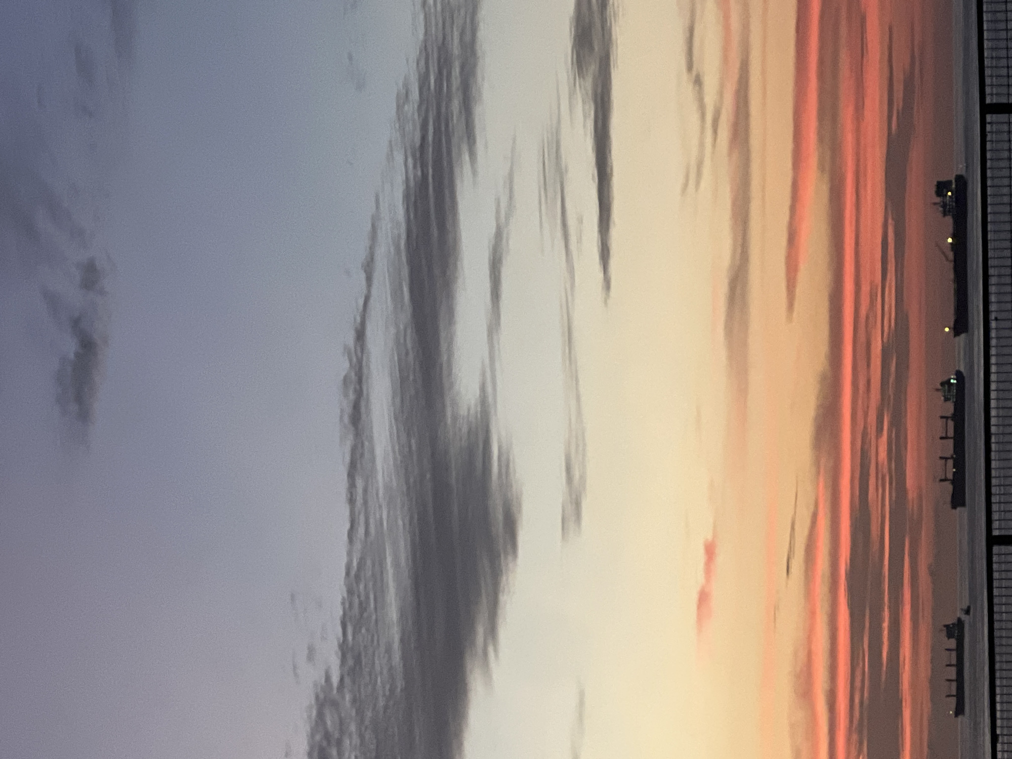 The image depicts a sunset sky over Arica, Chile. The sky transitions from blue-gray on the left to orange and red hues on the right. Streaky clouds are visible across the entire sky. Several ships are docked in the harbor. A fence-like structure is visible on the extreme right edge.