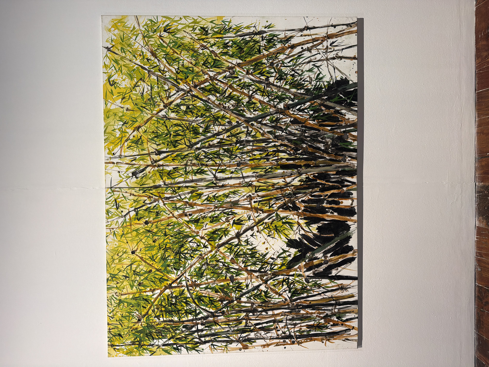 The image shows a tall, narrow painting on a white wall. The painting depicts a dense cluster of bamboo stalks and leaves rendered in a loose, expressive style. The bamboo is predominantly green and yellow, with brown and black accents for the stalks and shadows. The brushstrokes are visible and energetic, suggesting movement and a sense of natural growth. The composition is abstract, focusing on the textures and interplay of light and shadow within the bamboo thicket. There is no visible text in the image. The painting is hung in a gallery or art space, as indicated by the plain white wall and the adjacent wooden paneling, which may be part of the building's architecture or a display element. The lighting suggests an indoor environment, possibly with natural light supplementing artificial sources. The overall impression is one of organic vitality and a focus on the visual qualities of the bamboo itself. The location context of La Cuesta, Spain, does not provide specific visual cues within the image itself, but situates the artwork in a particular geographical and cultural setting.