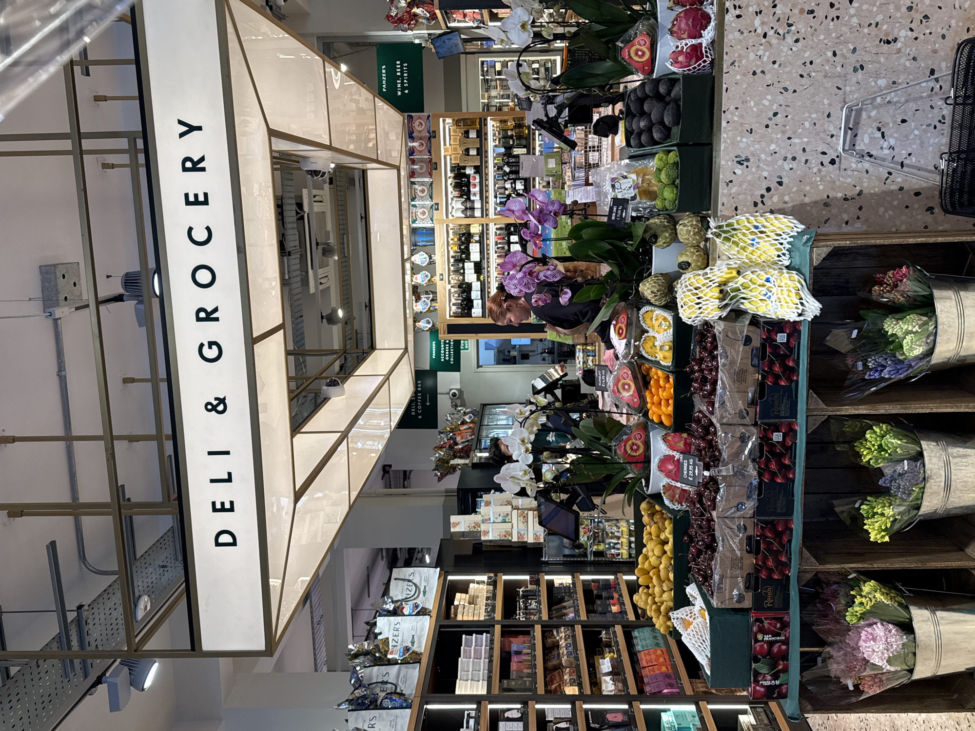 The image depicts the interior of a deli and grocery store identified as Panzer's, located in the St. John's Wood area of London, United Kingdom.
The scene features a brightly lit, modern retail space. In the foreground, a substantial display table is laden with fresh produce including yellow citrus fruits, red cherries in labeled boxes (£29.95/kg), dragon fruit, kumquats, cherimoyas, kaffir limes (£75.00/kg), avocados, and bananas in mesh bags. Potted purple and white orchids are interspersed among the produce. Below this table, bouquets of hyacinths and other flowers are arranged in silver buckets within wooden crates. A black metal shopping basket rests on the terrazzo-patterned floor to the right.
In the midground, a woman with red hair, wearing a dark top, stands behind the main counter, facing away from the camera. Partially visible further back is another individual in a green uniform. Behind the counters, shelves are stocked with various packaged goods, including gourmet items, and bottles of wine, beer, and spirits. Overhead, a large illuminated sign reads "DELI & GROCERY" in black capital letters. Additional green signs indicate "PANZER'S DELI, SANDWICHES & COFFEE BAR," "PANZER'S WINE, BEER & SPIRITS," and "PANZER'S ACCOMPANIMENTS & GIFTS COLLECTION." The ceiling features exposed ductwork, cable trays, and track lighting. The overall environment suggests an upscale grocery establishment.