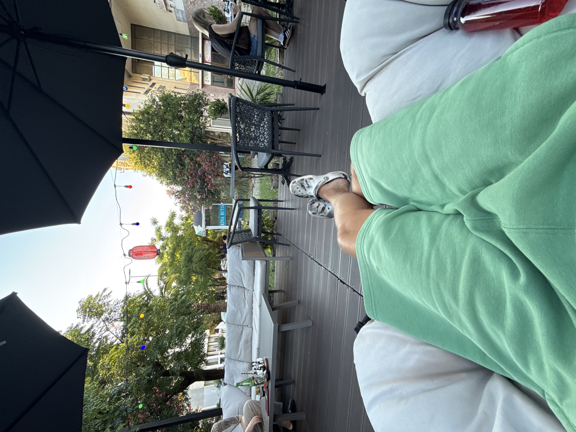 The image depicts an outdoor seating area of a cafe or restaurant, possibly in Bar, Montenegro, given the visual context of palm trees and Mediterranean-style architecture in the background. In the foreground, a person's legs are visible, resting on a white cushion. They are wearing light green shorts and grey patterned Crocs. The perspective suggests the photo was taken from a seated position, looking out at the scene.
The seating area is furnished with grey wooden decking, black umbrellas providing shade, and outdoor furniture. A large, light grey sectional sofa is visible to the left, with a grey coffee table in front of it. On the table are a bottle of water, a straw, and a small decorative sailboat. Further into the scene, there are black woven chairs and small tables.
In the background, a building with large windows and some signage is present. Among the foliage, there are decorative string lights with colorful bulbs and a red paper lantern. A woman with blonde hair is seated at a table to the right, wearing a dark top and denim shorts, and appears to be engaged with her phone. The overall atmosphere is relaxed and casual, suggesting a daytime setting.