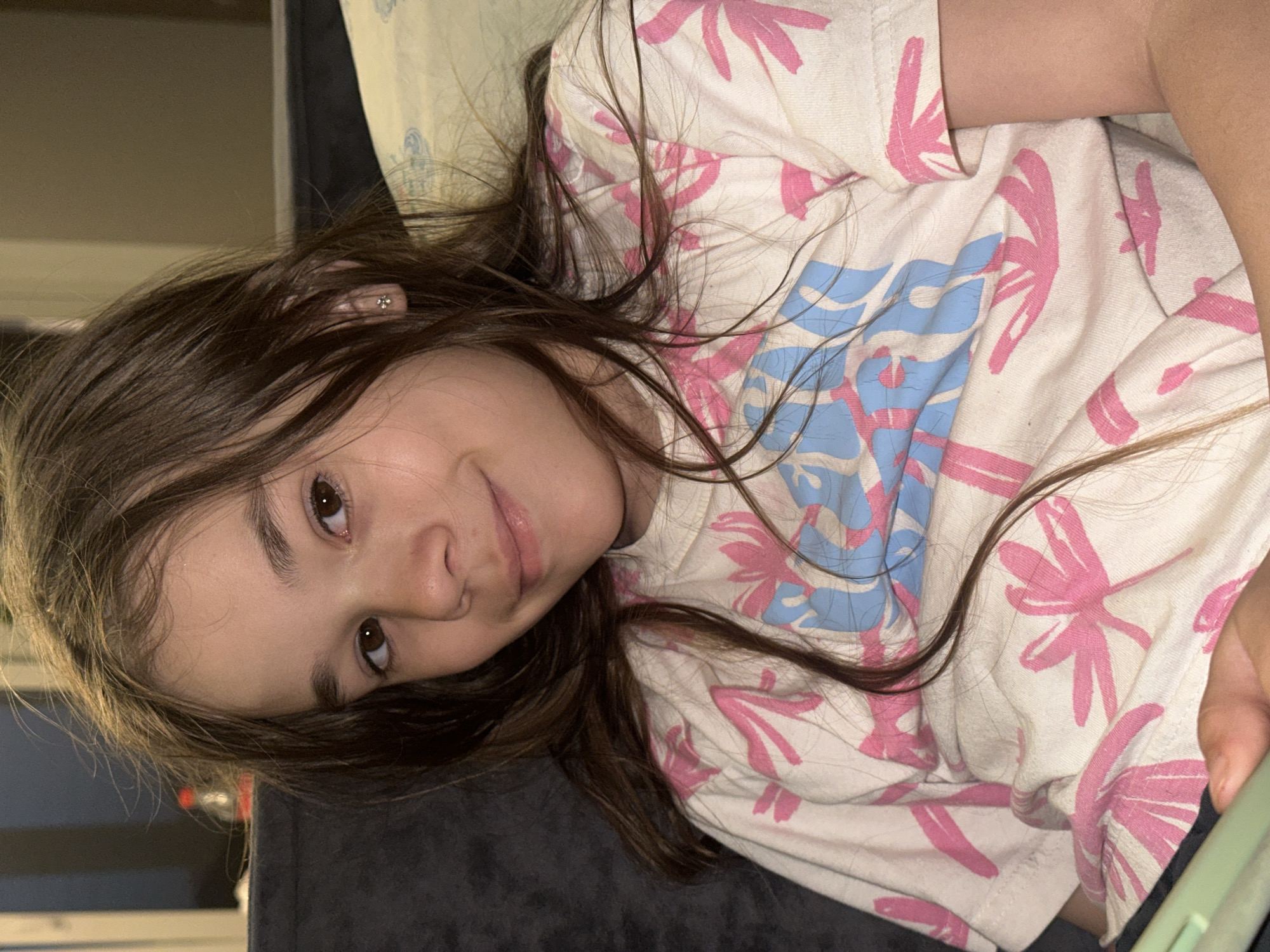 A young girl with long, brown hair is lying down, resting her head on a dark gray surface. She is wearing a white t-shirt with a pink floral and blue wave pattern. She has an earring in her left ear. There is no indication of the city of Kharnavy, Russia, or any specific location. The scene appears to be indoors.
