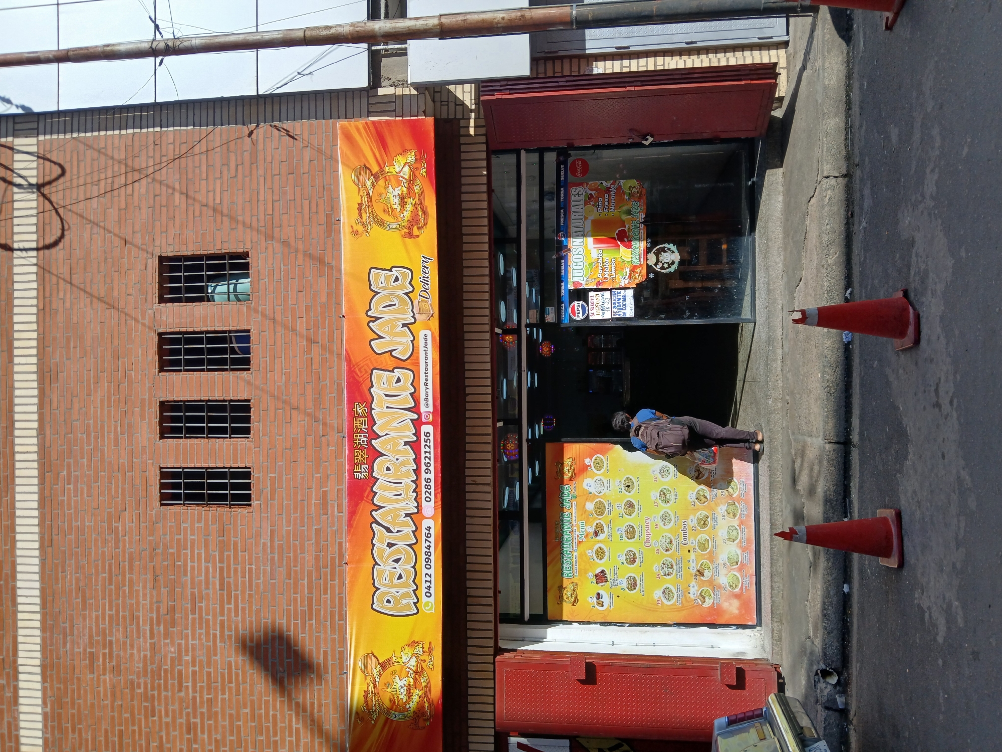 A street-level view shows the exterior of a building in Ciudad Guayana, Venezuela, that houses a Chinese restaurant called Restaurante Jade. The building has a brick facade, with a vertical banner displaying the restaurant's name and contact information. There are three barred windows on the left side of the brick facade. The restaurant's entrance is visible, flanked by menus and posters. An individual is entering the restaurant. Two orange traffic cones are placed on the sidewalk in front of the restaurant.