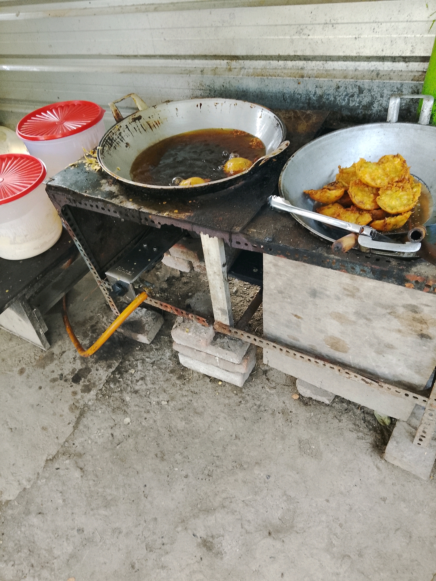 In Banaran, Indonesia, a scene of street food preparation unfolds. Two aluminum pans sit atop a makeshift cooking station constructed from scrap metal and brick supports. The left pan contains bubbling oil, with a few fritters immersed, while the right pan holds a collection of golden-brown fried items, likely a local delicacy. A metal tong rests on the edge of the right pan, suggesting the food is either being removed or placed for draining. Two white plastic containers with red lids are visible to the left, potentially used for storing ingredients or finished products. The cooking station is situated outdoors, with a concrete ground surface bearing stains and debris, indicative of a typical street-side operation. The background features a corrugated metal wall, common in such informal settings. An orange gas hose snakes from beneath the cooking station, connecting to a gas source that fuels the cooking.