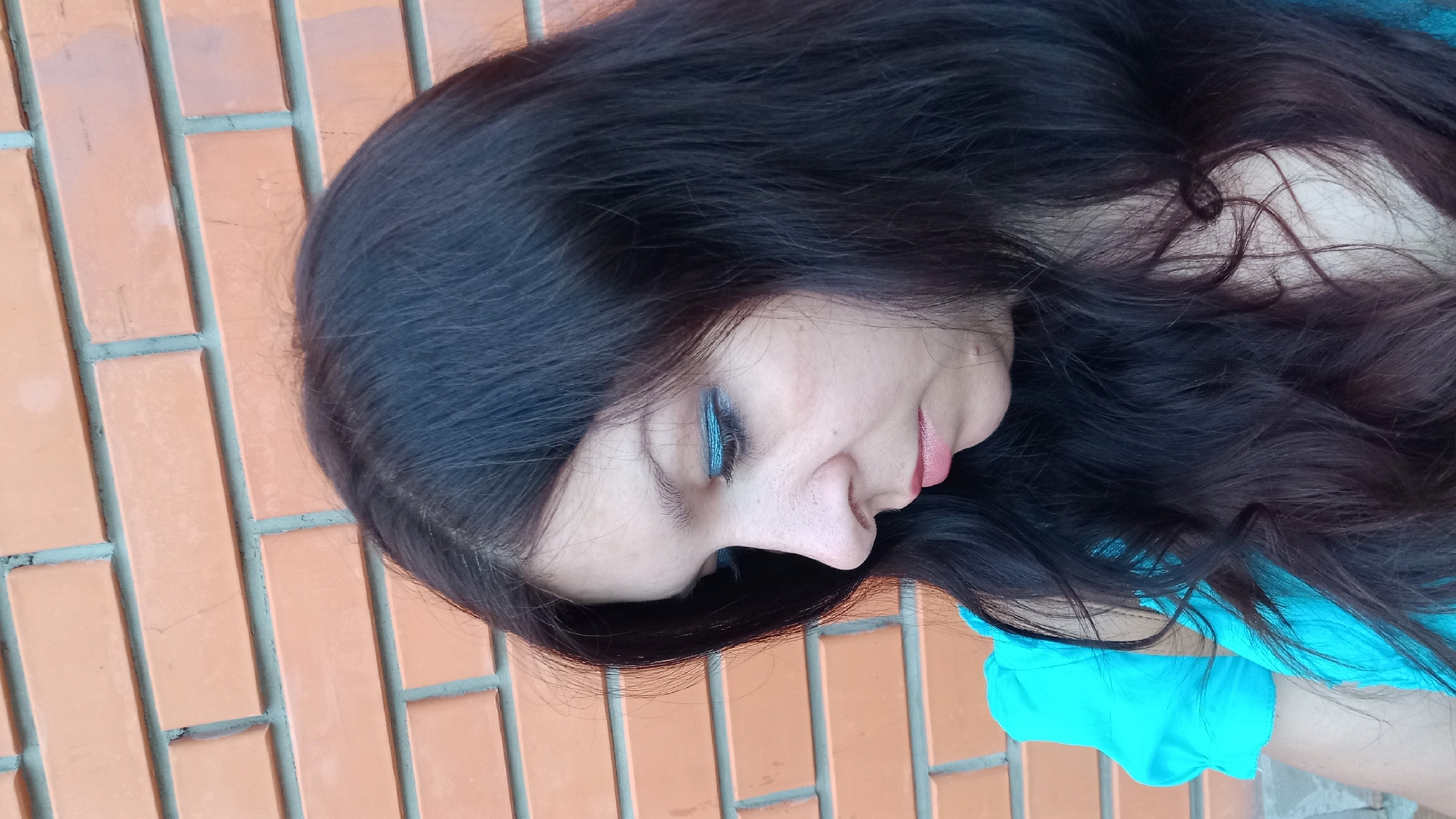 A medium shot profile of a woman with long, dark hair, wearing a turquoise top, stands against a brick wall. She is looking down and to her right, with blue eyeshadow on her eyelids. The brick wall is a pale orange with grey mortar. The location is Kropyvnytskyi, Ukraine.