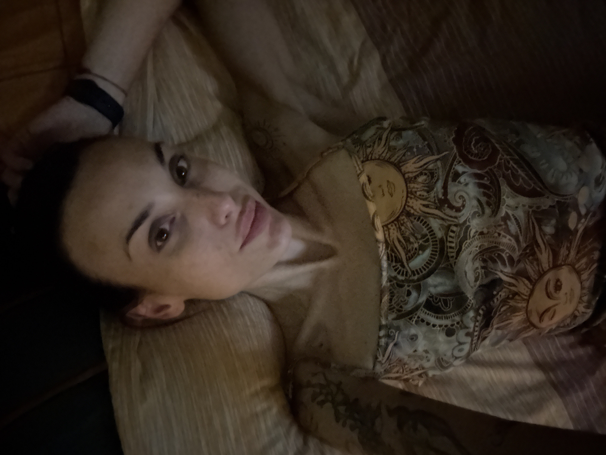 The image shows a close-up, high-angle, overhead shot of a woman lying on a bed. She is looking at the camera with a slight smile. Her head is tilted back, and her left arm is raised above her head, with her hand resting on the back of her head. She is wearing a tank top with a sun and paisley pattern. There are tattoos visible on her left arm and shoulder. The background is a textured bedspread. The image was taken indoors, likely in a bedroom. There is no information in the image to suggest a location in Pleven, Bulgaria.